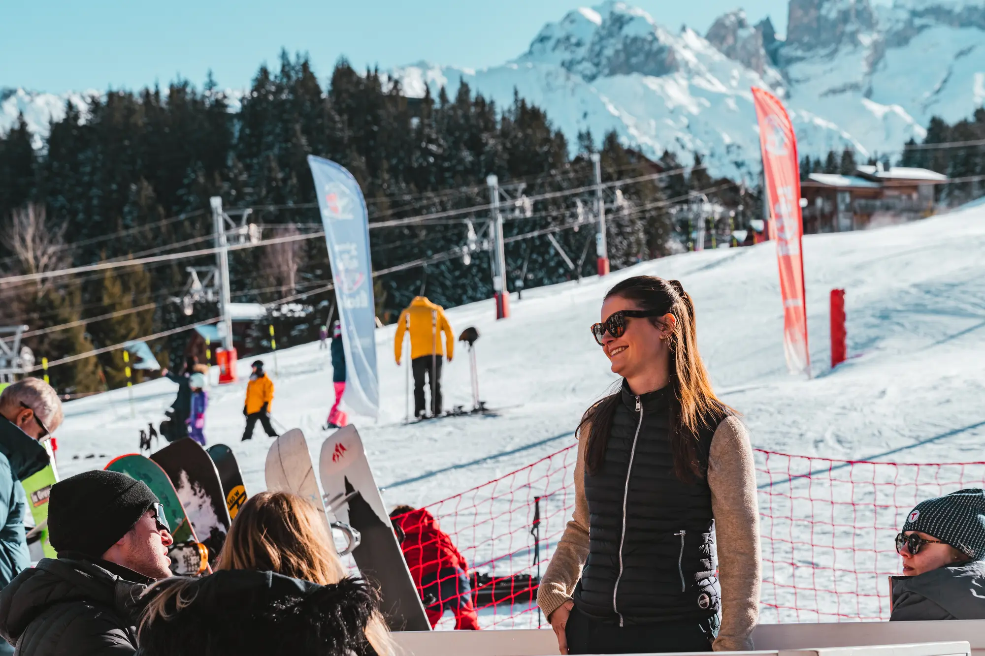 Une femme souriante portant des lunettes de soleil et un gilet noir discute avec deux personnes assises sur un banc au pied d'une piste de ski enneigée, avec des skieurs, des snowboards, un filet de sécurité rouge, des sapins et des montagnes enneigées en arrière-plan.