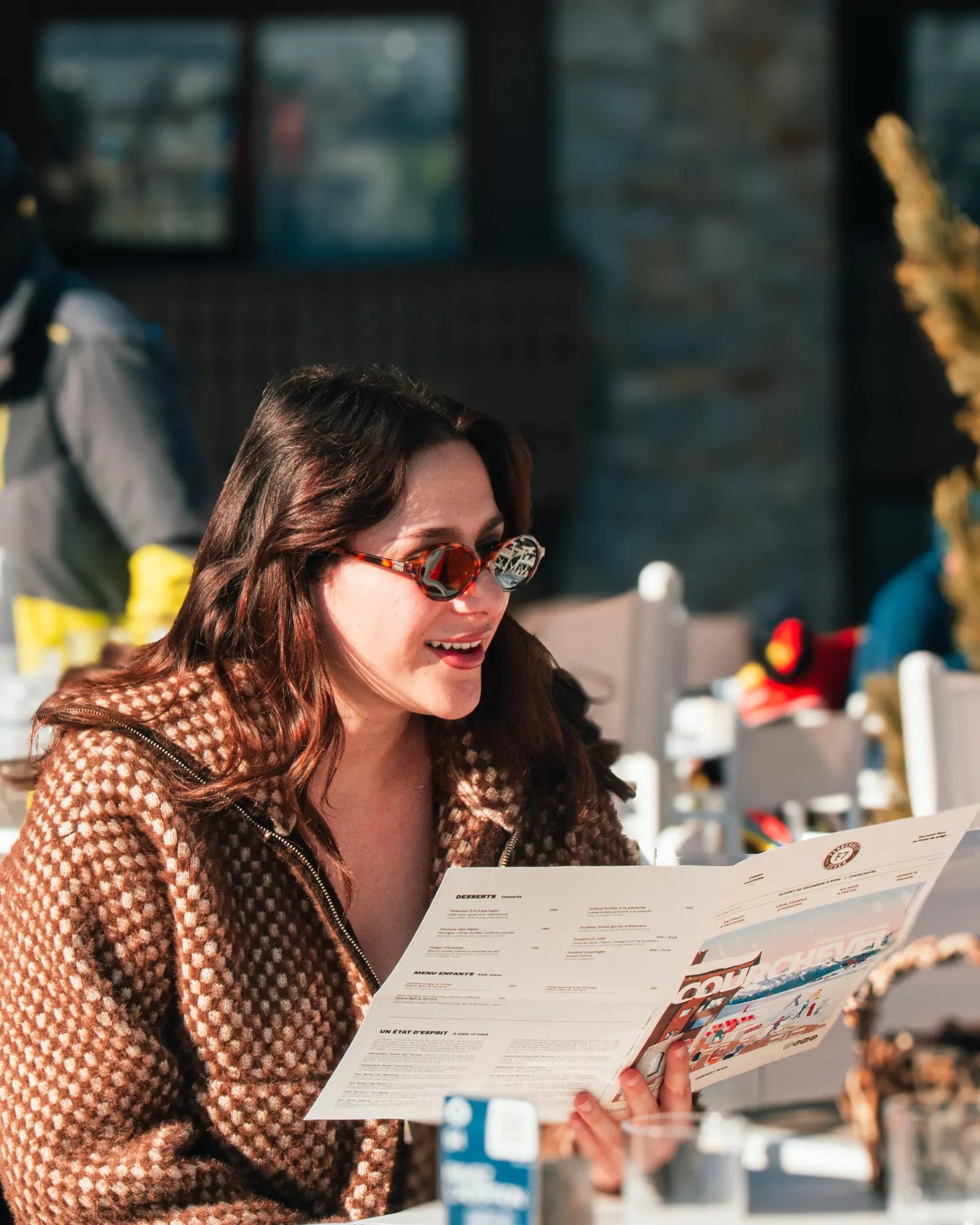 Une femme portant des lunettes de soleil rétro et un manteau à motifs marron et beige tient un menu de restaurant en souriant, assise à une table extérieure ensoleillée.