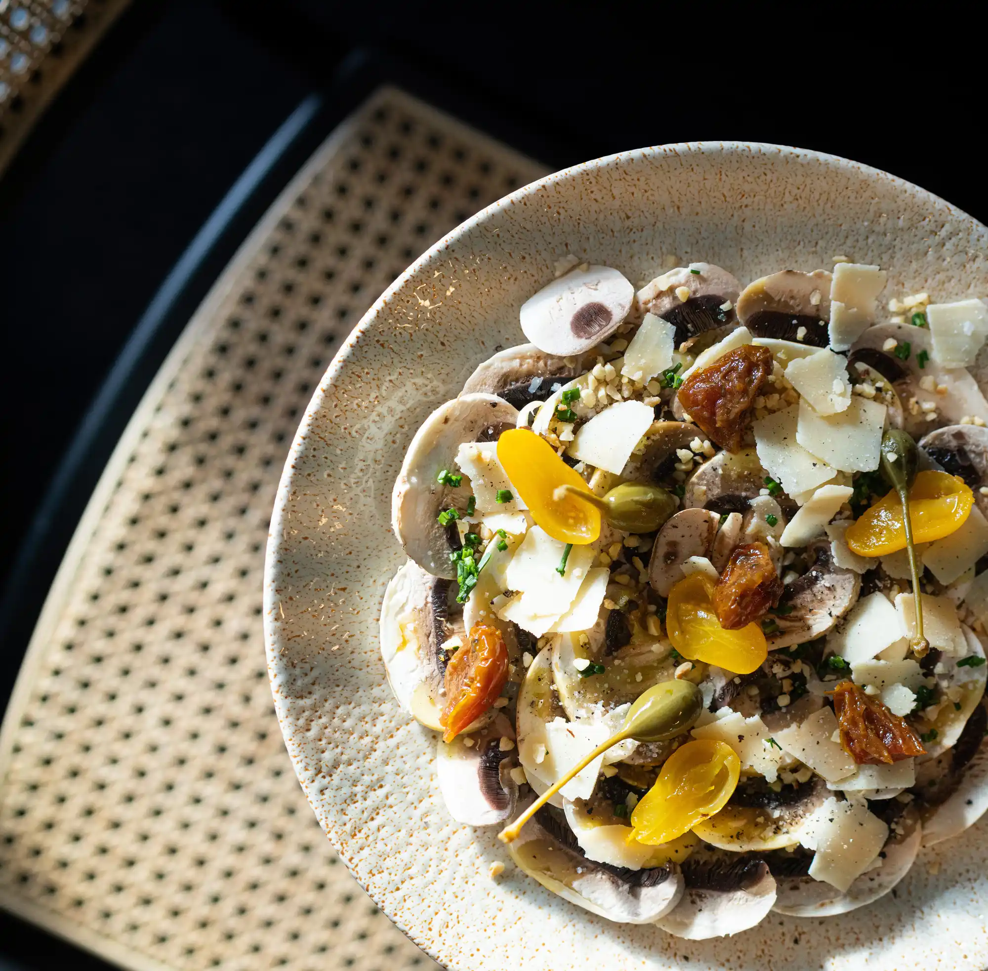Assiette de salade composée de fines tranches de champignons frais, copeaux de fromage, câpres, raisins secs dorés et herbes fraîches, posée sur une surface en osier tressé.