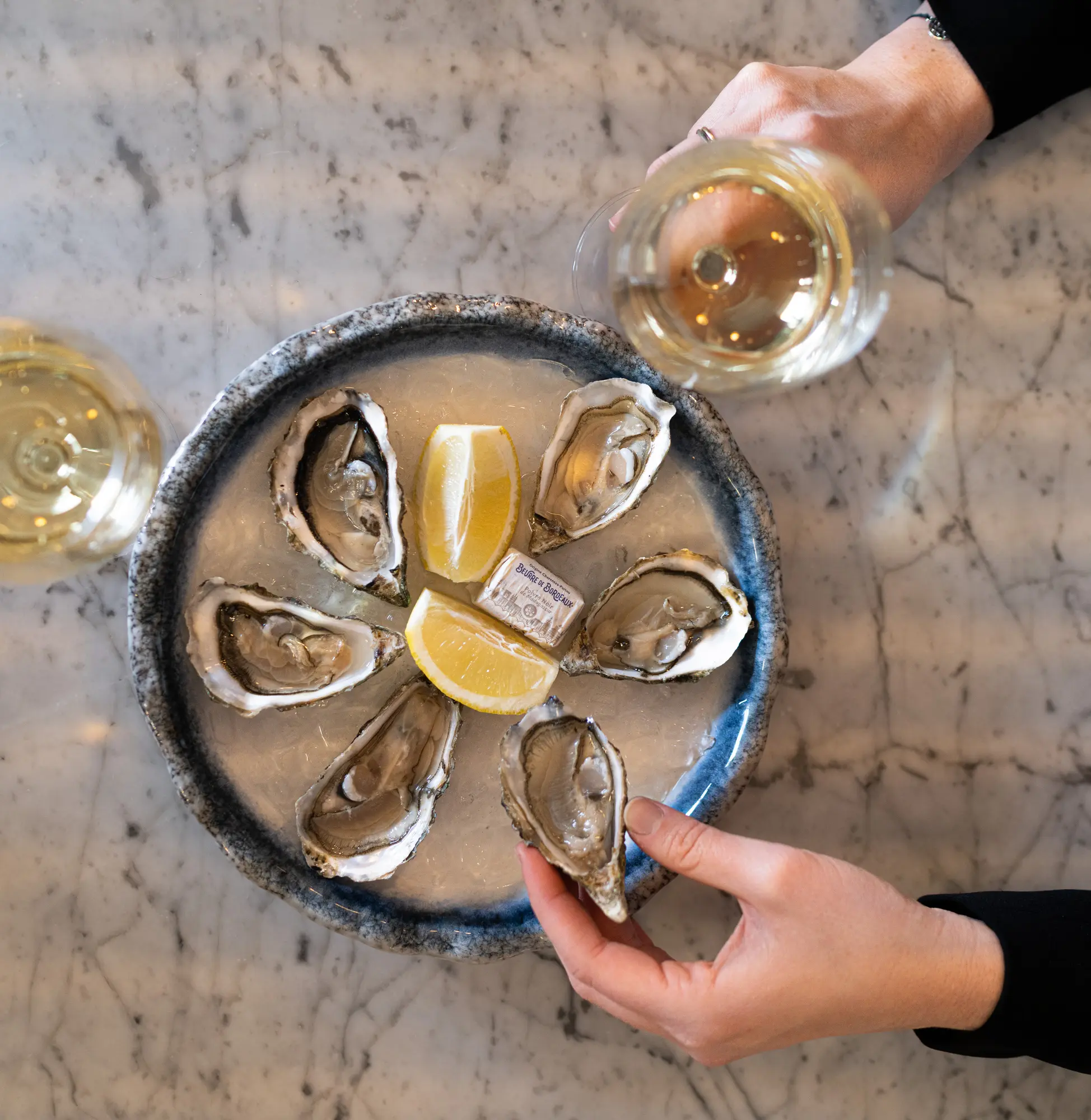 Une assiette ronde avec six huîtres ouvertes sur un lit de glace, garnie de quartiers de citron et d'un petit paquet de beurre, posée sur une table en marbre avec deux verres de vin blanc et des mains tenant une huître et un verre.