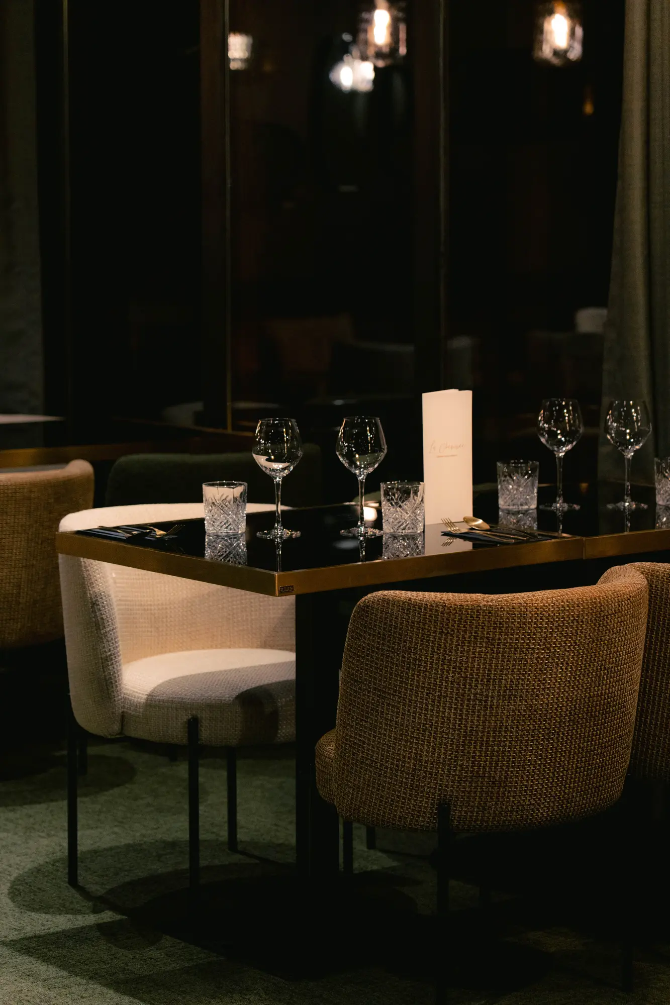 Table élégamment dressée avec quatre verres à vin, quatre verres à eau en cristal, des couverts et un menu debout, entourée de fauteuils en tissu beige et marron dans un restaurant au décor sombre.
