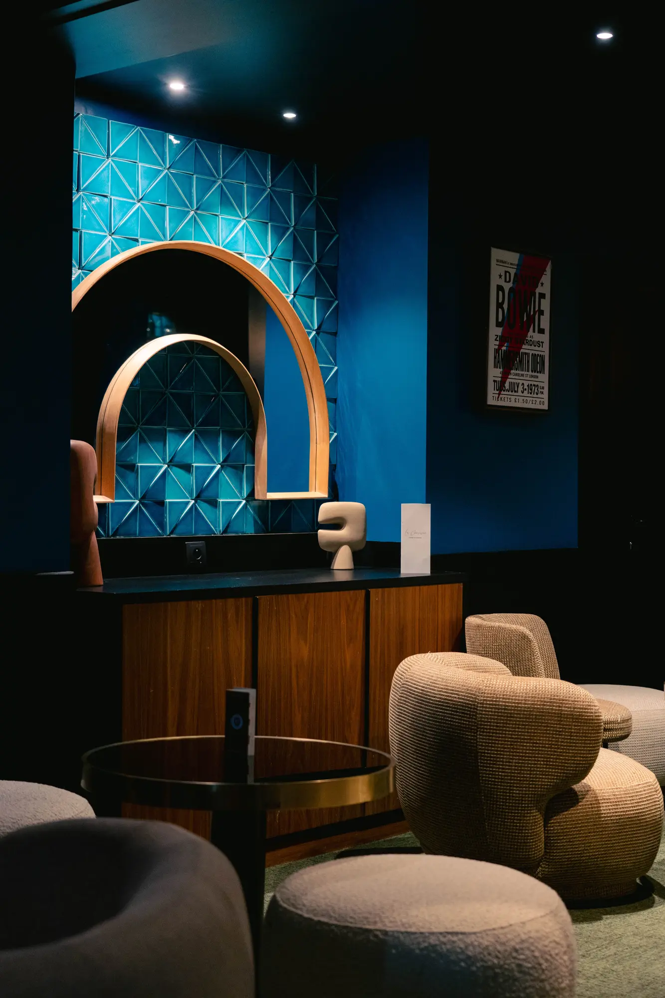 Intérieur moderne avec un mur bleu orné de carreaux texturés et un miroir en bois en forme d'arche, une commode en bois foncé avec une petite sculpture abstraite, une chaise beige rembourrée et une table ronde avec un plateau en verre foncé.