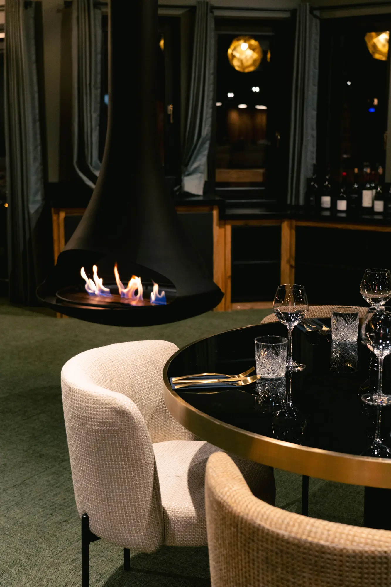 Coin repas élégant avec une table ronde noire entourée de chaises beige en tissu, un foyer suspendu avec des flammes visibles, et des verres à vin posés sur la table dans une ambiance chaleureuse.