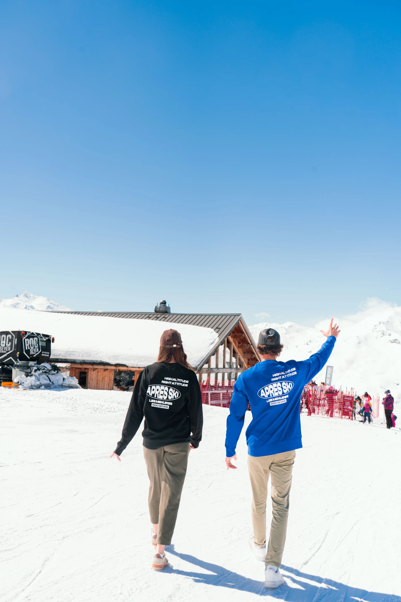 Deux personnes marchant sur la neige vers un bâtiment en bois avec un toit métallique partiellement couvert de neige, portant des sweats à capuche avec l'inscription Apres Ski et d'autres mots, sous un ciel bleu clair et ensoleillé dans une station de ski avec des montagnes enneigées en arrière-plan.