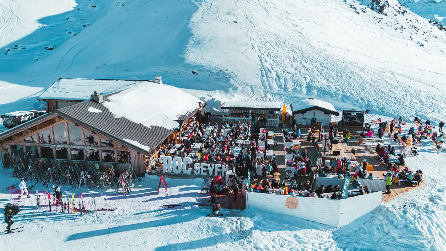 Terrasse extérieure d'un restaurant de montagne nommé ROC SEVEN avec beaucoup de personnes assises et socialisant sur des tables et des chaises en bois, entourée de neige avec des skis appuyés contre le bâtiment.