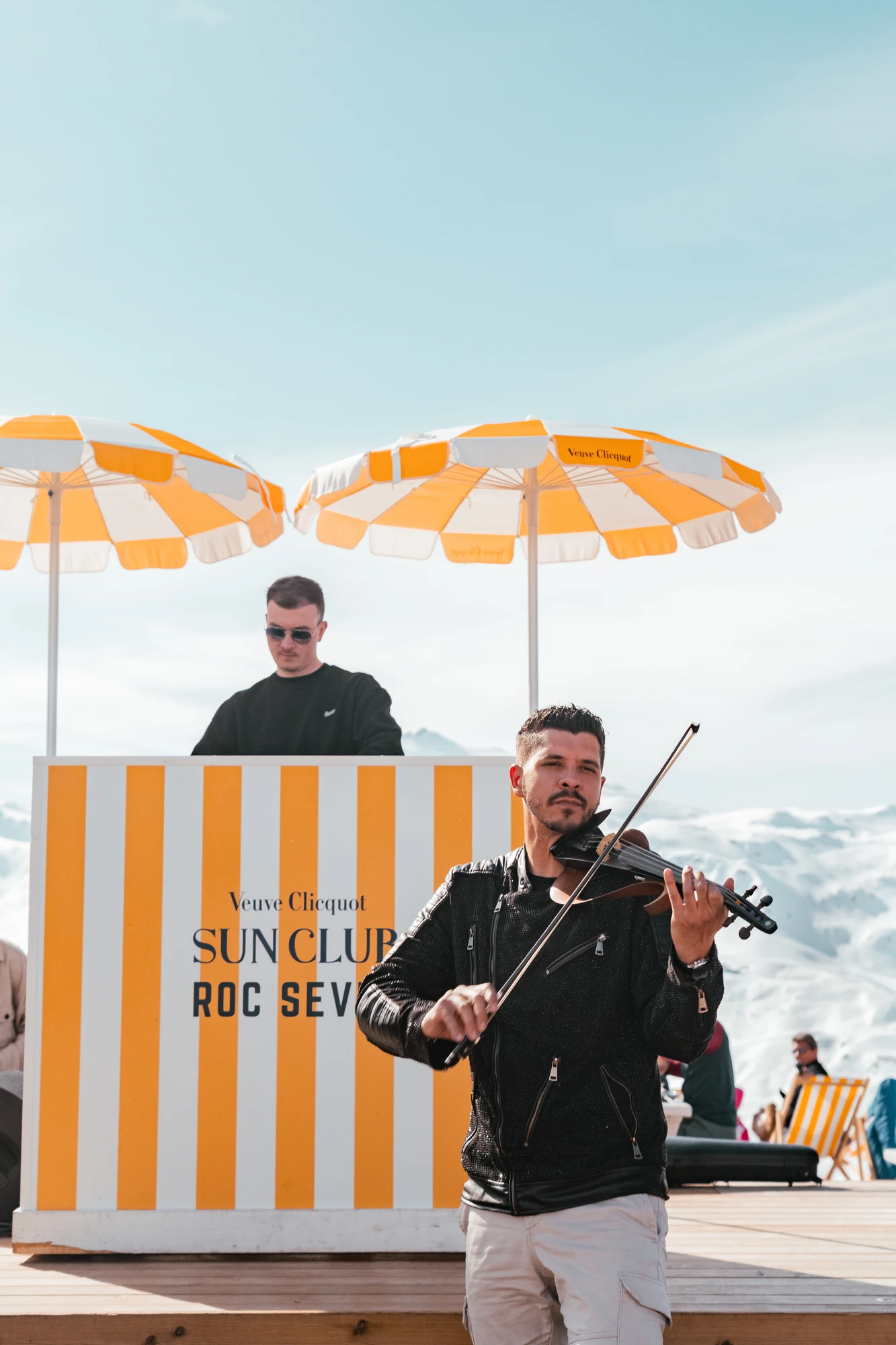 Un homme jouant du violon devant un stand rayé jaune et blanc avec le texte Veuve Clicquot SUN CLUB ROC SEV, sous un ciel clair, avec un autre homme portant des lunettes de soleil debout derrière le stand, deux parasols jaunes et blancs au-dessus, et des montagnes enneigées en arrière-plan.