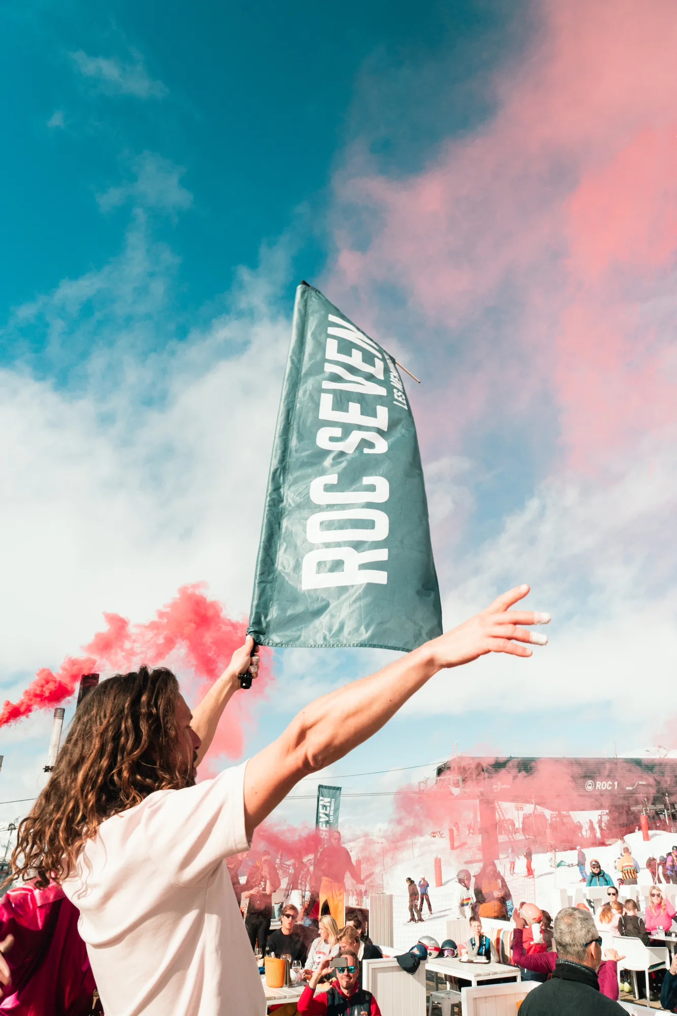 Person avec les cheveux longs tenant un drapeau marqué "ROC SEVEN" en plein air, entouré de fumée rouge et de gens assis et debout sous un ciel bleu avec des nuages.