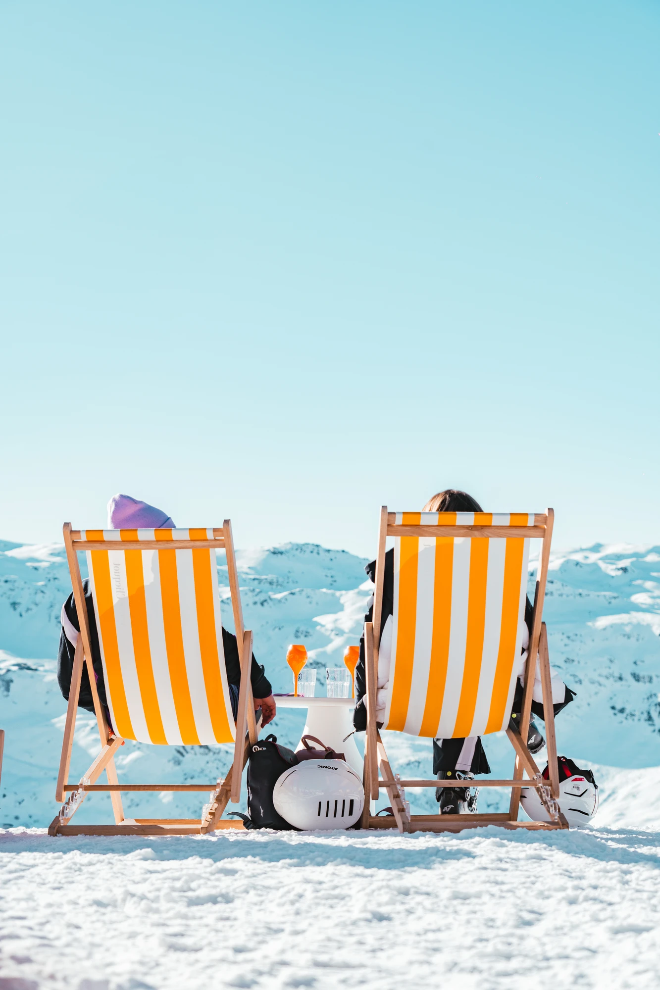 Deux personnes assises sur des chaises longues à rayures jaunes et blanches, face à des montagnes enneigées sous un ciel clair, avec deux verres orange posés sur une table entre elles.