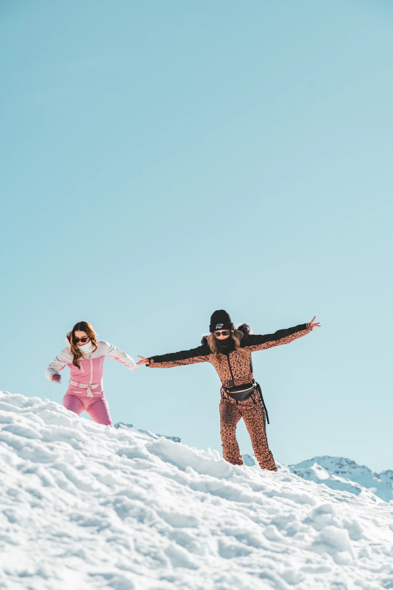 Deux femmes habillées en combinaisons de ski colorées, tenant la main et posant de façon ludique sur une pente enneigée avec un ciel bleu clair et des montagnes en arrière-plan.