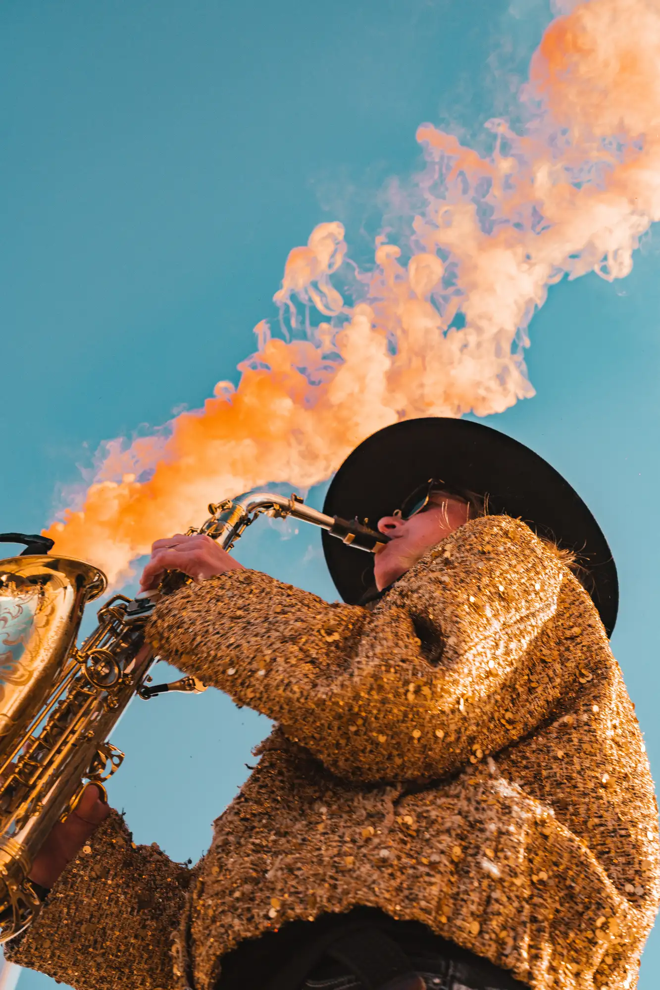 Personne portant un chapeau noir et un manteau doré avec des sequins jouant du saxophone sous un ciel bleu clair, avec de la fumée orange sortant de l'instrument.