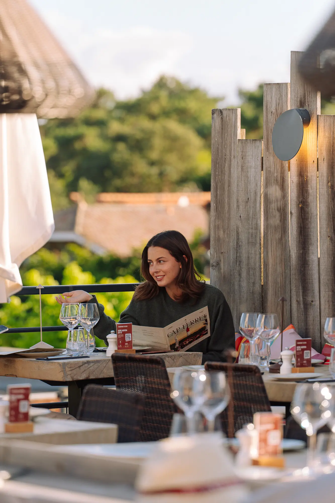 Une femme souriante assise seule à une table extérieure dans un restaurant, tenant un menu Cap Ferret, avec plusieurs verres à vin vides sur la table et un décor en bois en arrière-plan.