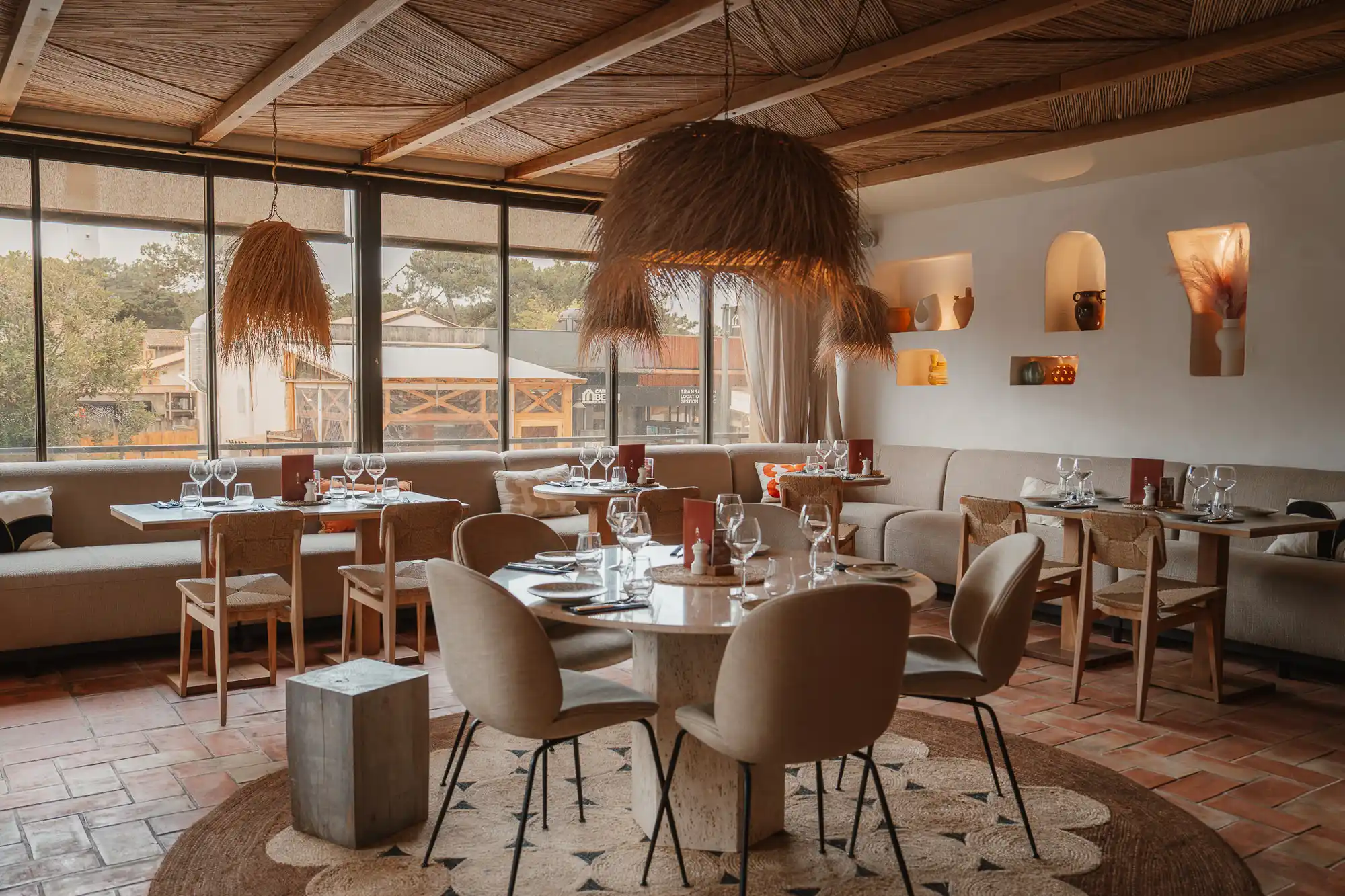 Intérieur d'un restaurant avec grandes fenêtres, tables dressées de verres et assiettes, chaises en bois et rembourrées, luminaires en fibres naturelles suspendus et décoration murale avec niches éclairées.