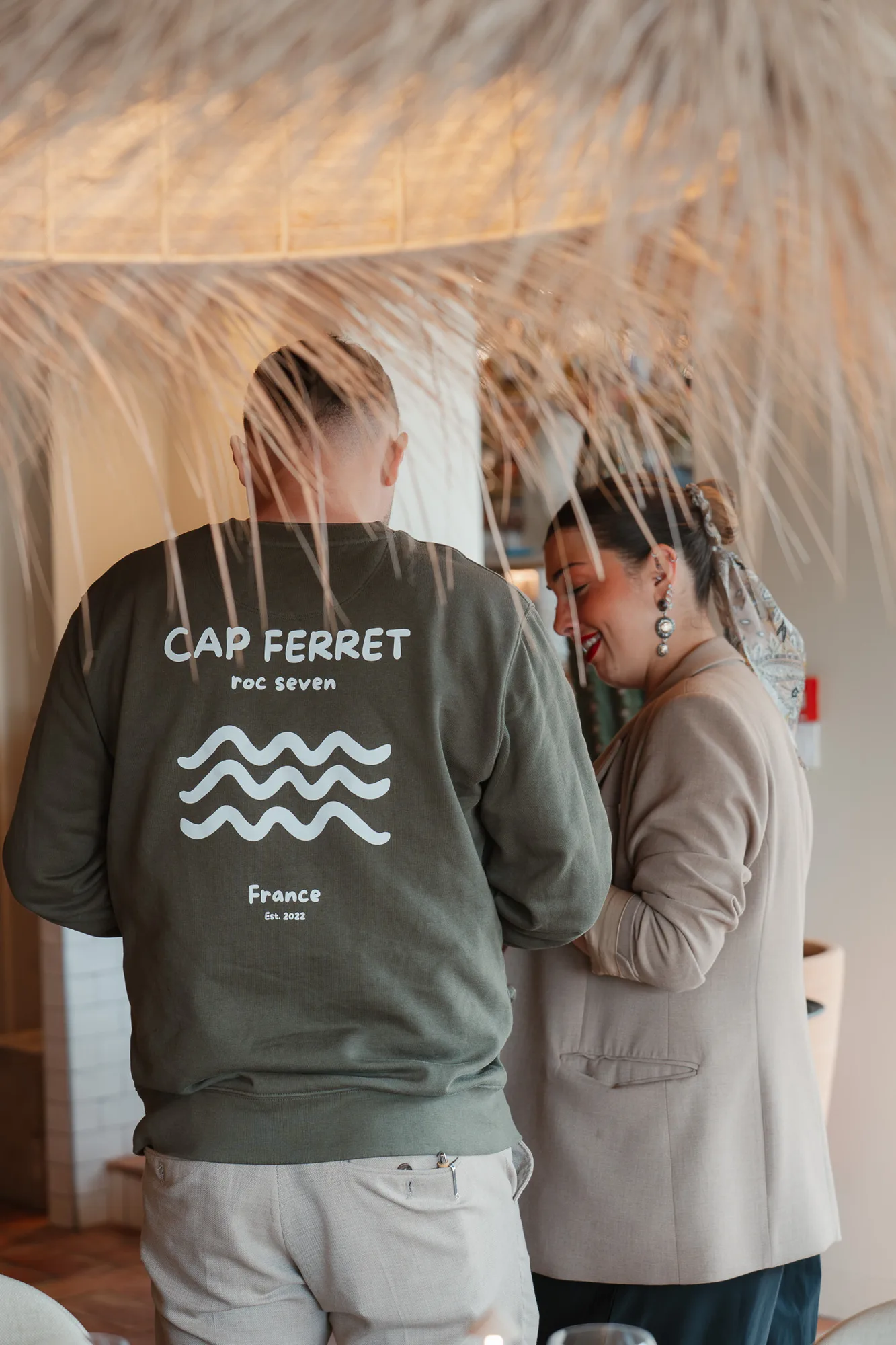 Un homme de dos portant un sweat vert olive avec l'inscription blanche « CAP FERRET roc seven France Est. 2022 » et un motif de vagues, à côté d'une femme souriante en blazer beige avec un foulard dans les cheveux et de grandes boucles d'oreilles, dans un cadre intérieur avec une décoration naturelle en premier plan.