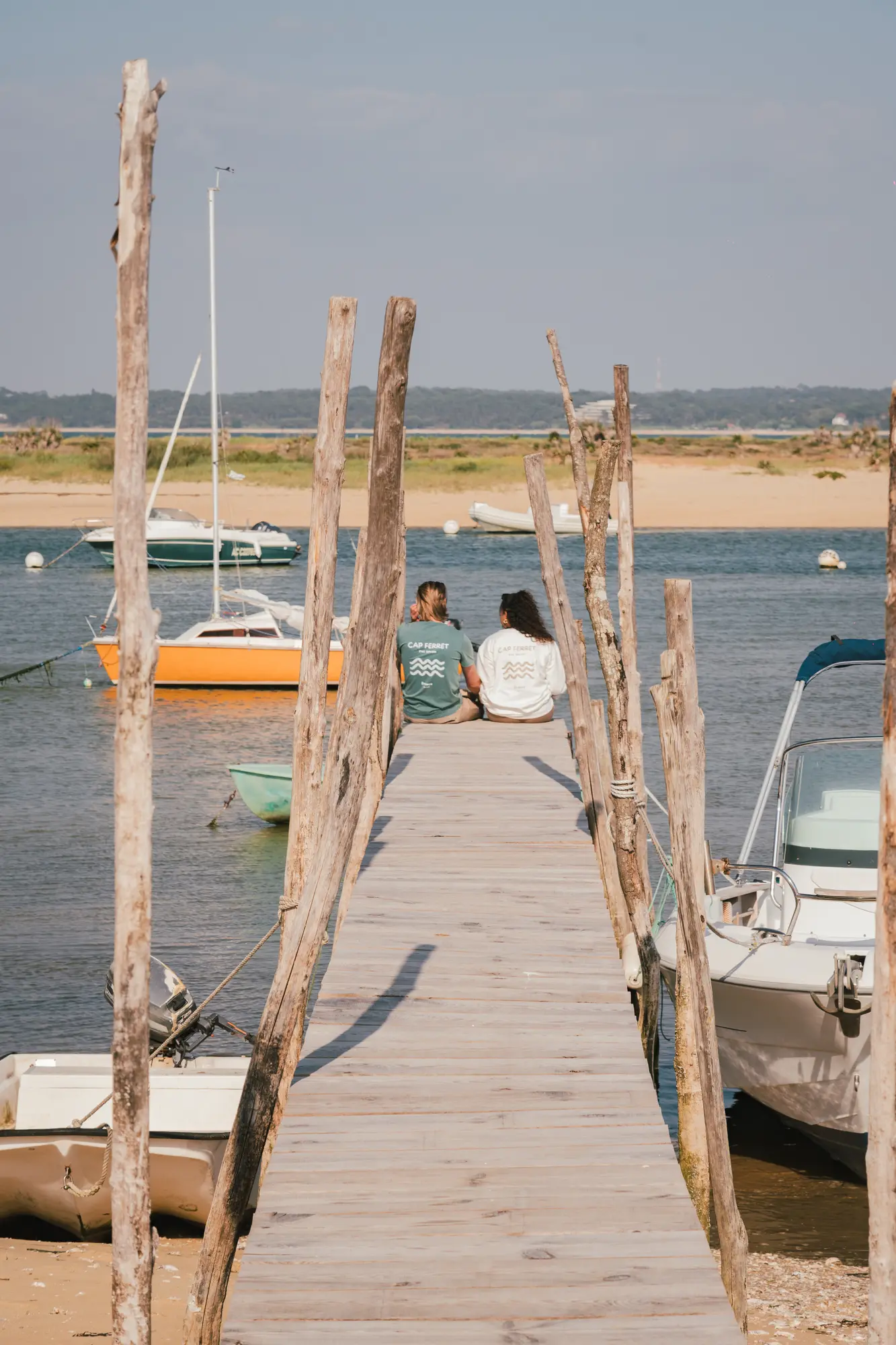 Deux personnes assises au bout d'une jetée en bois avec des poteaux en bois, face à un plan d'eau avec des bateaux amarrés et une plage en arrière-plan.