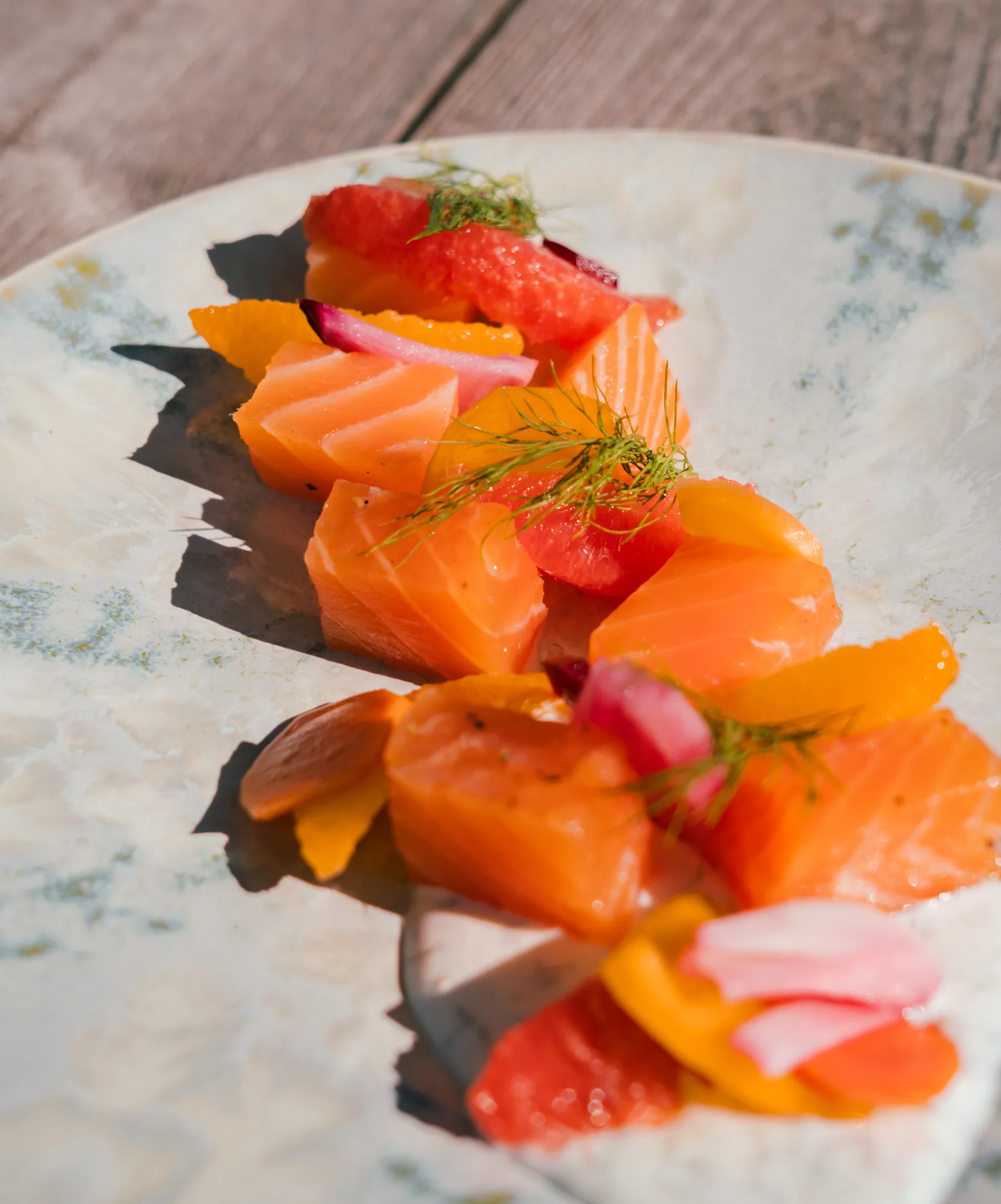 Morceaux de saumon cru joliment présentés sur une assiette avec des segments d'agrumes colorés, des tranches d'oignon rouge et des herbes vertes.