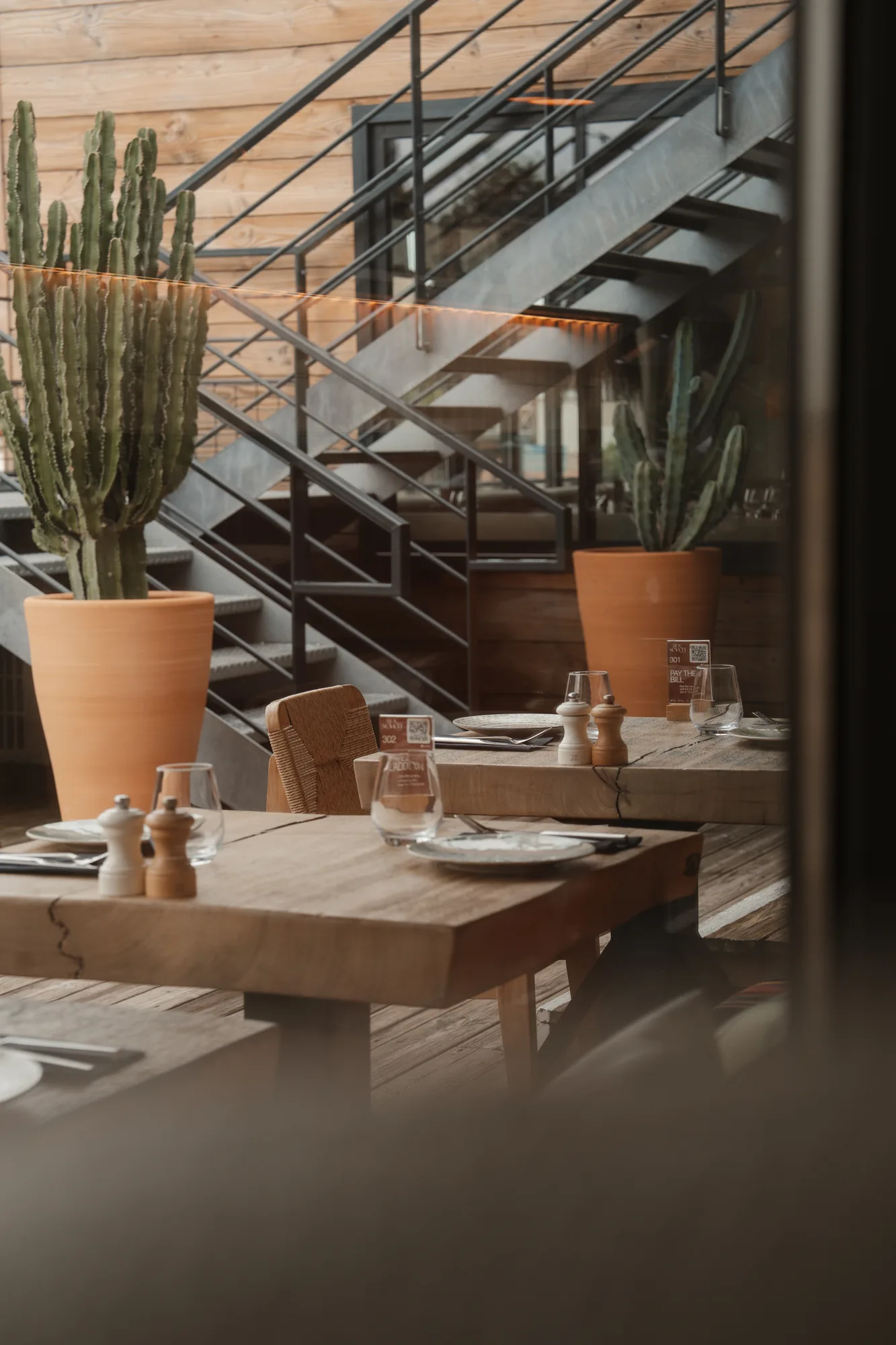 Intérieur d'un restaurant avec tables en bois, chaises tressées, grandes plantes cactus en pots, et un escalier en métal noir à l'arrière.