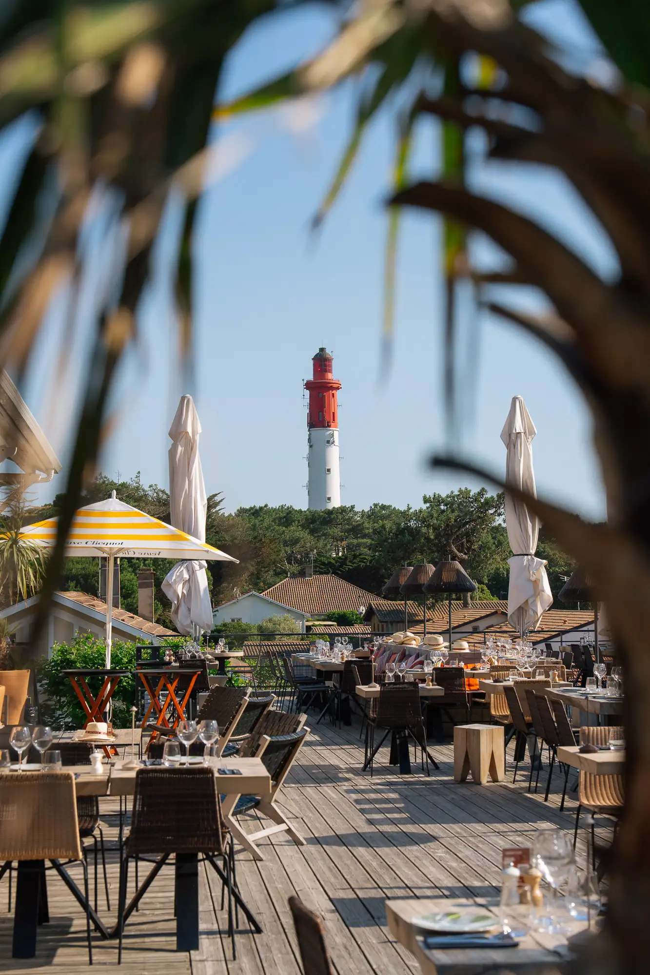 Terrasse de restaurant en bois avec tables et chaises dressées, parasols fermés et un parasol rayé jaune et blanc, vue encadrée par des feuilles, avec un phare rouge et blanc en arrière-plan sous un ciel bleu clair.