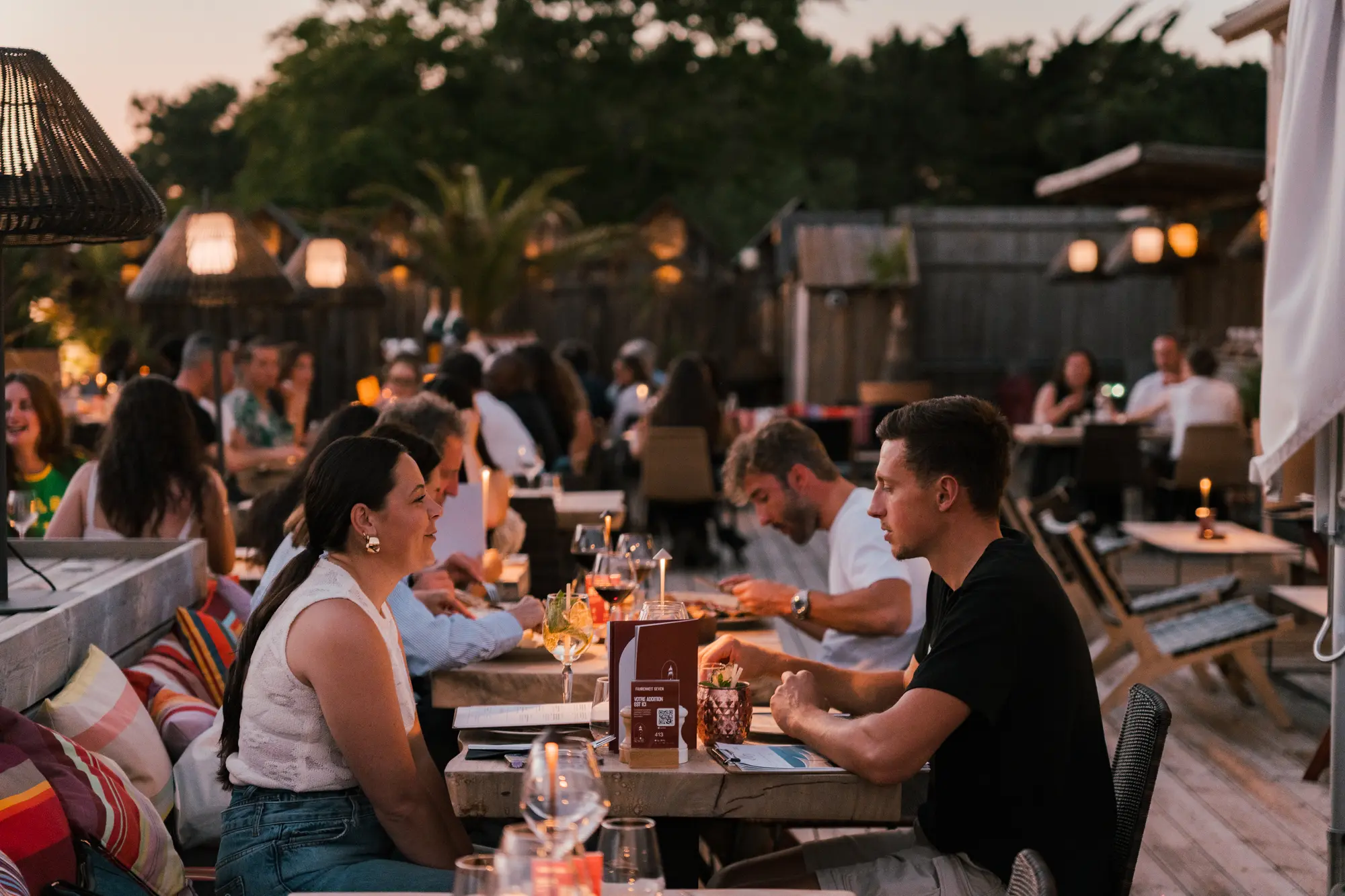 Dîner en plein air au crépuscule dans un restaurant animé avec des personnes assises à une table en bois, discutant et dégustant des boissons et des repas.