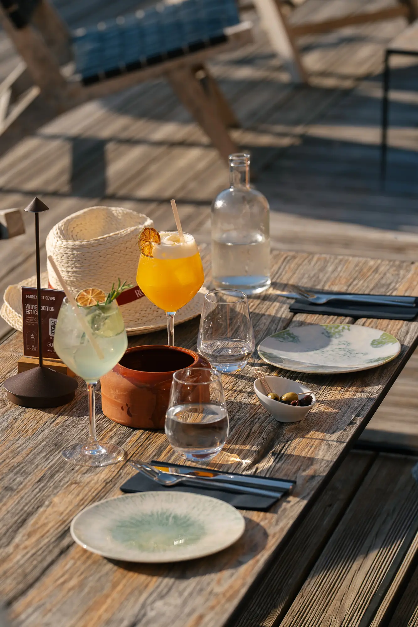 Table en bois extérieure avec deux assiettes, deux verres d'eau, deux cocktails colorés garnis d'agrumes, un petit bol d'olives, une carafe d'eau, une assiette en terre cuite ronde et un chapeau en paille.
