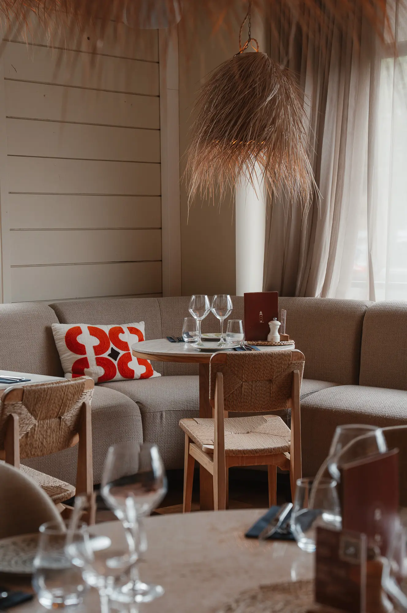 Coin repas élégant avec une banquette beige en forme de U, une table ronde avec verres à vin, assiettes, menu, chaises en bois tissé et une grande suspension en fibre naturelle suspendue au plafond, le tout dans une ambiance chaleureuse avec rideaux clairs.