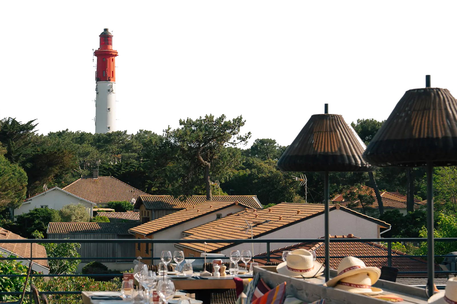 Vue extérieure d'une terrasse de restaurant avec des tables dressées, des verres et des chapeaux posés, en arrière-plan des toits de maisons et un phare blanc et rouge entouré d'arbres verts.
