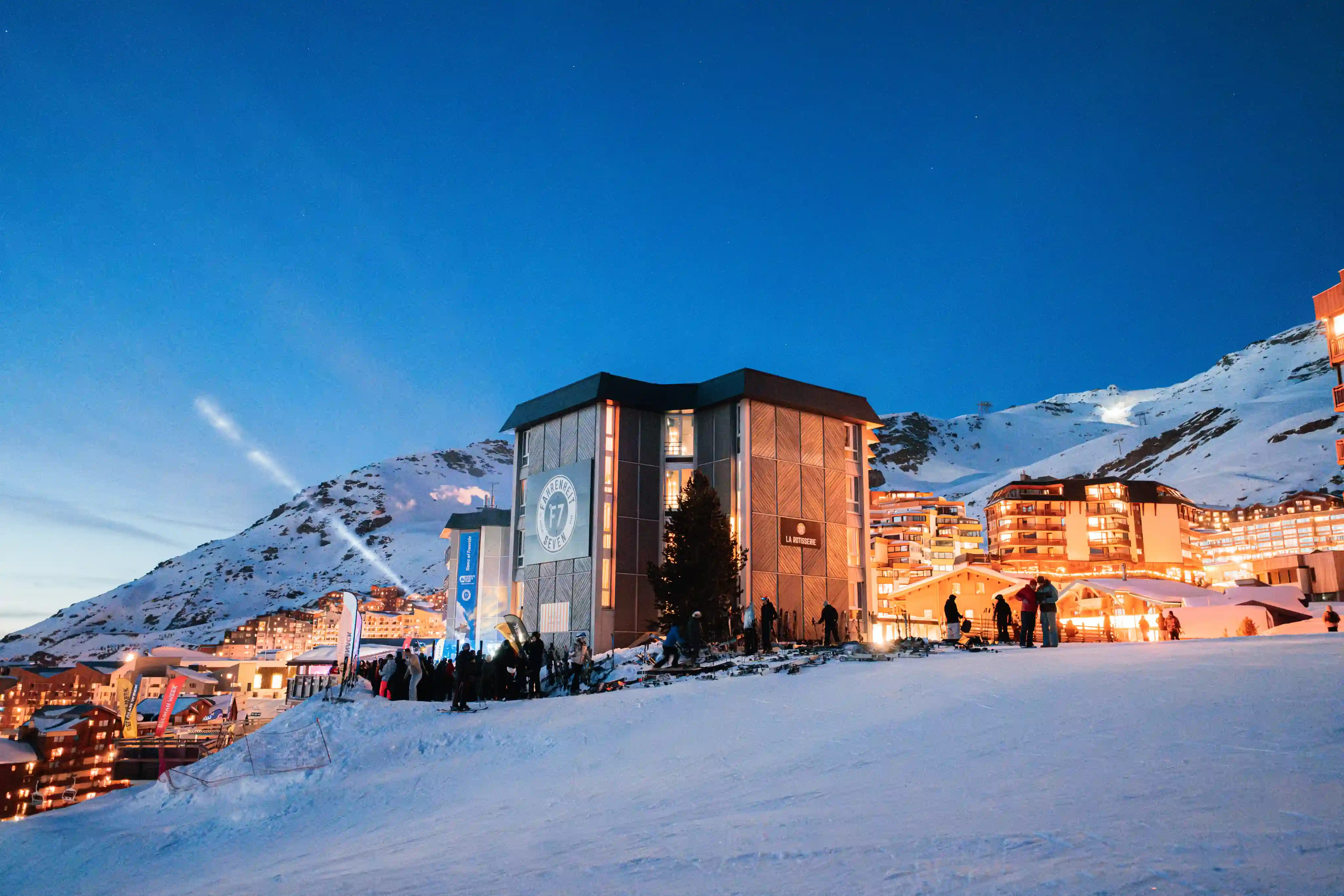 Station de ski au crépuscule avec un bâtiment marqué F7 entouré de skieurs, des montagnes enneigées en arrière-plan, et des lumières chaudes des bâtiments.