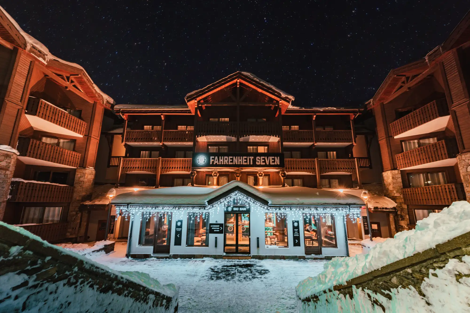 Hôtel illuminé appelé Fahrenheit Seven avec un bar de ski au premier plan, entouré de neige sous un ciel étoilé nocturne.