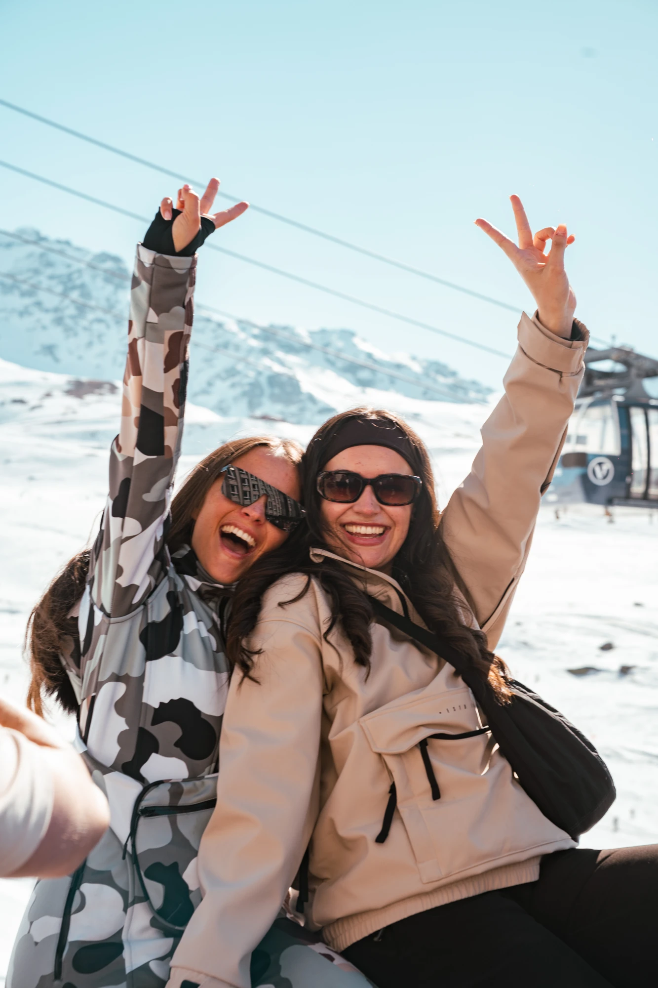 Deux femmes souriantes portant des lunettes de soleil et des vestes d'hiver, levant les bras avec des signes de paix, devant un paysage de montagne enneigée et une télécabine.