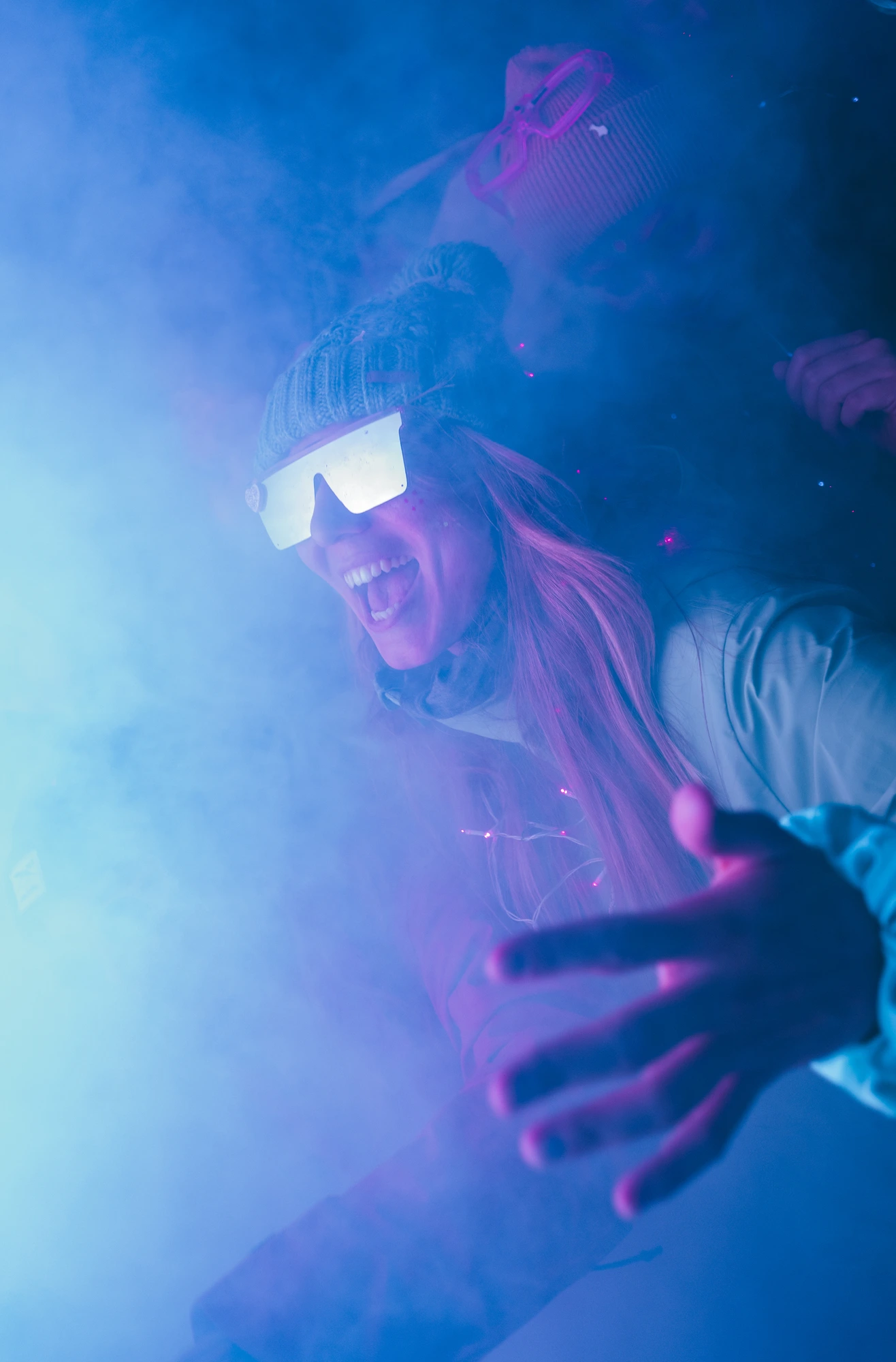 Deux personnes portant des lunettes de protection futuristes et des bonnets en tricot dans une atmosphère enfumée avec une lumière bleue et violet, l'une souriant largement et tendant la main vers l'objectif.
