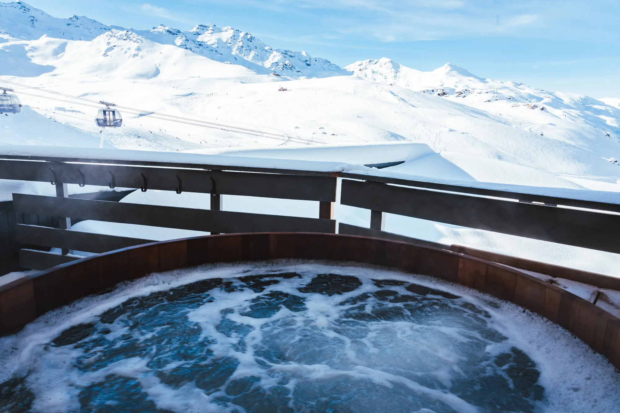 Bain à remous en bois sur un balcon avec vue sur des montagnes enneigées et des télécabines sous un ciel bleu clair.