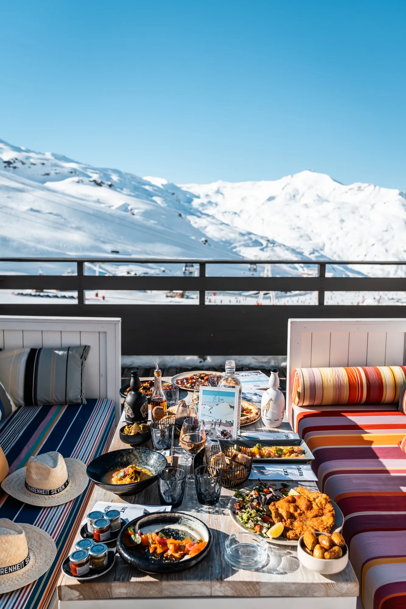 Table extérieure dans une station de ski avec une variété de plats, dont pizzas, salade, pommes de terre et boissons, entourée de banquettes colorées et vue sur les montagnes enneigées sous ciel bleu.