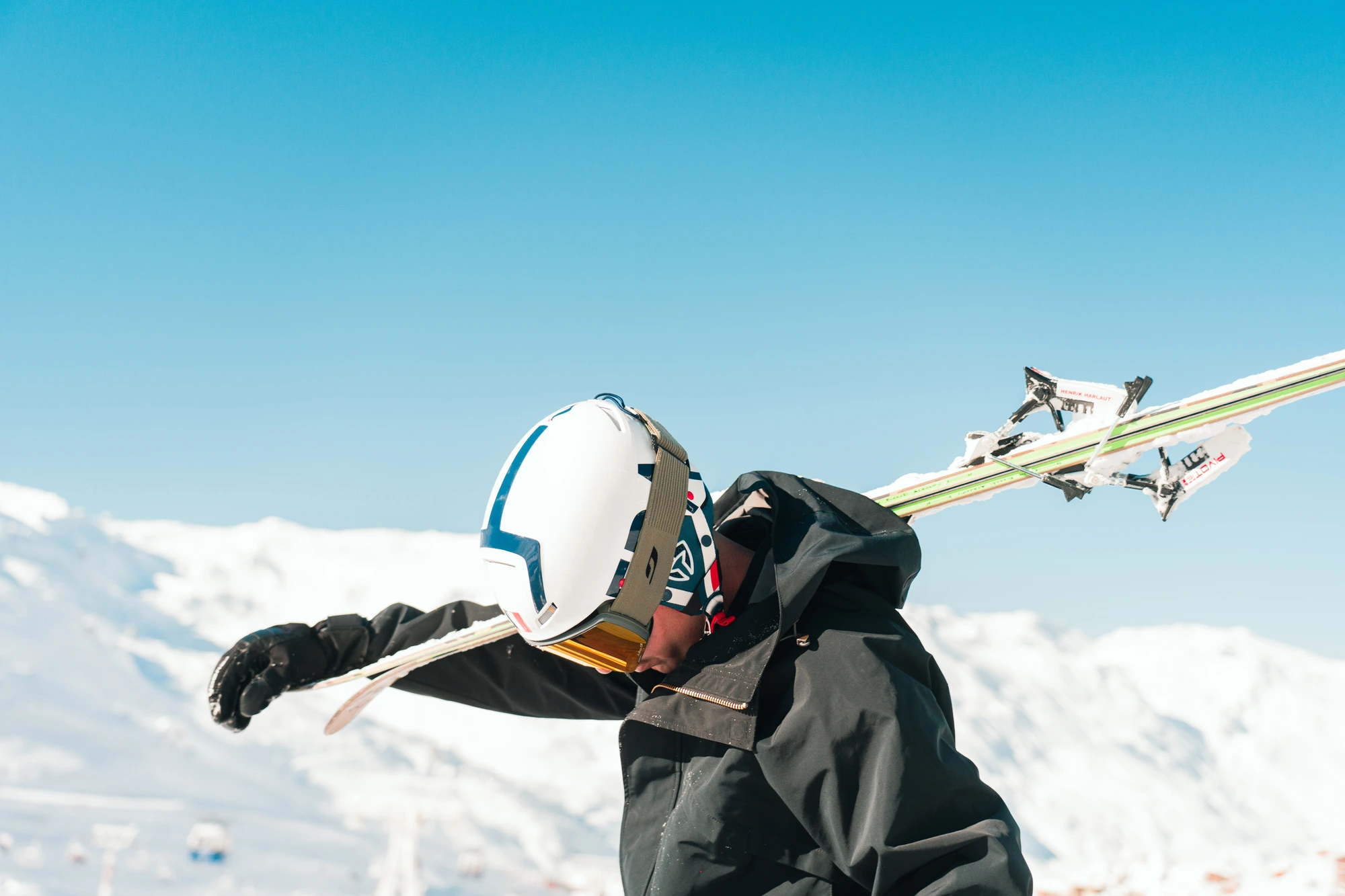 Un skieur portant un casque blanc et des lunettes jaunes, habillé d'une veste noire, porte ses skis sur son épaule dans un paysage de montagne enneigée sous un ciel bleu clair.