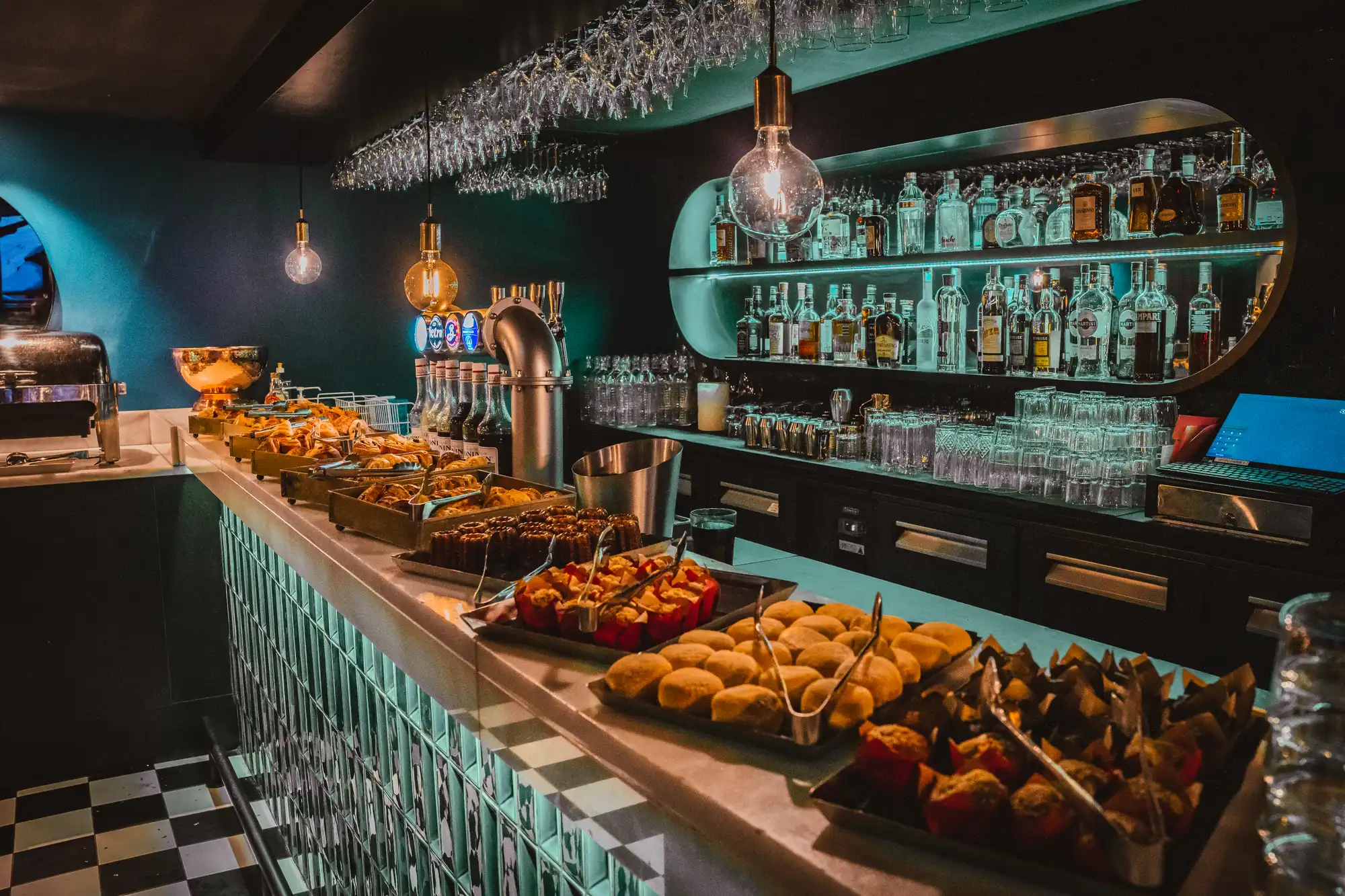 Bar avec un comptoir présentant divers plateaux de pâtisseries et petites viennoiseries, éclairé par des ampoules suspendues, avec des étagères de bouteilles d'alcool en arrière-plan et un ordinateur portable ouvert sur le côté.