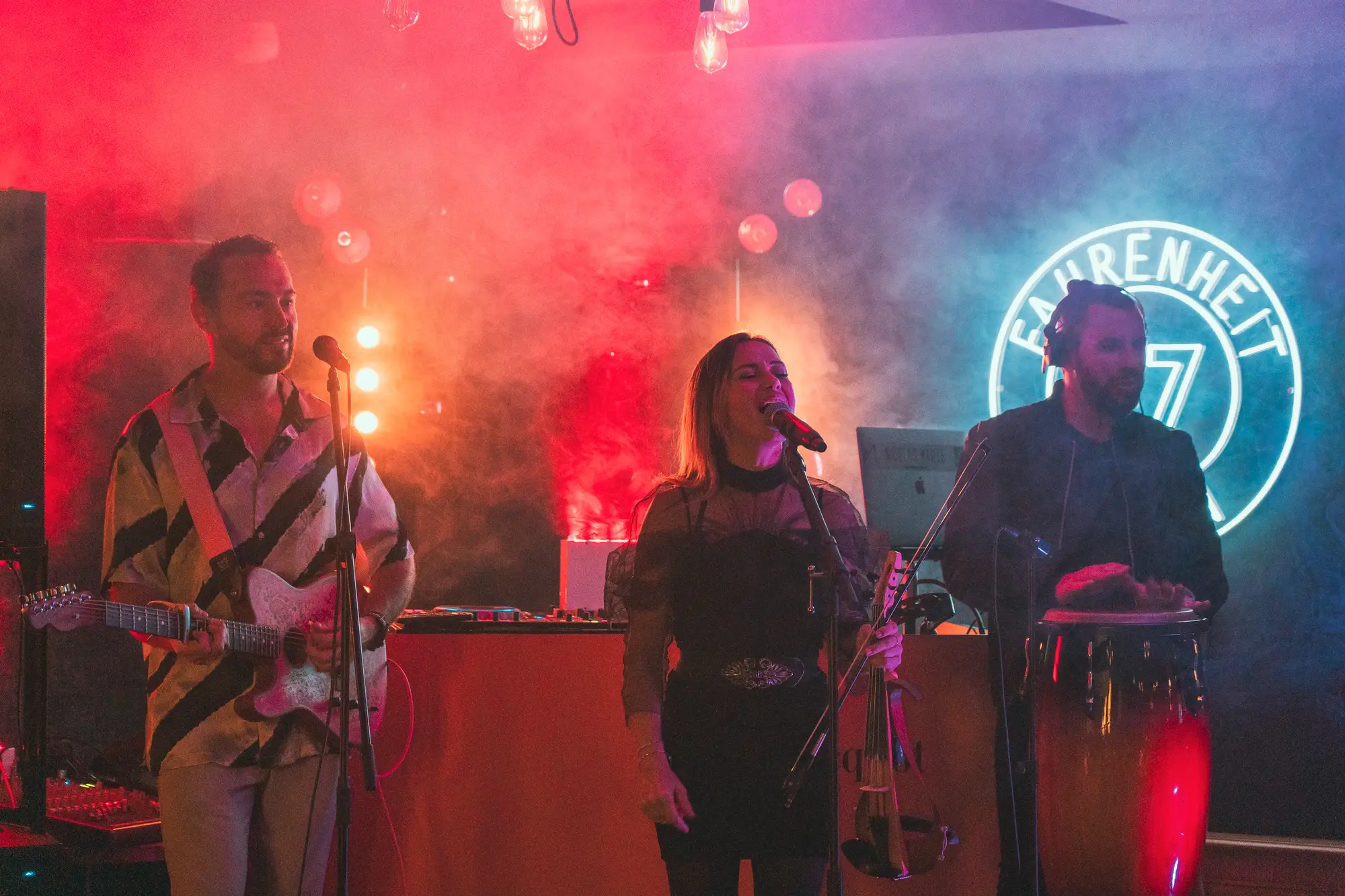 Un groupe de musiciens sur scène avec un éclairage rouge et bleu, un homme joue de la guitare électrique, une femme chante au micro avec un violon électrique, et un homme joue des congas à droite sous un logo lumineux « Fahrenheit 7 ».