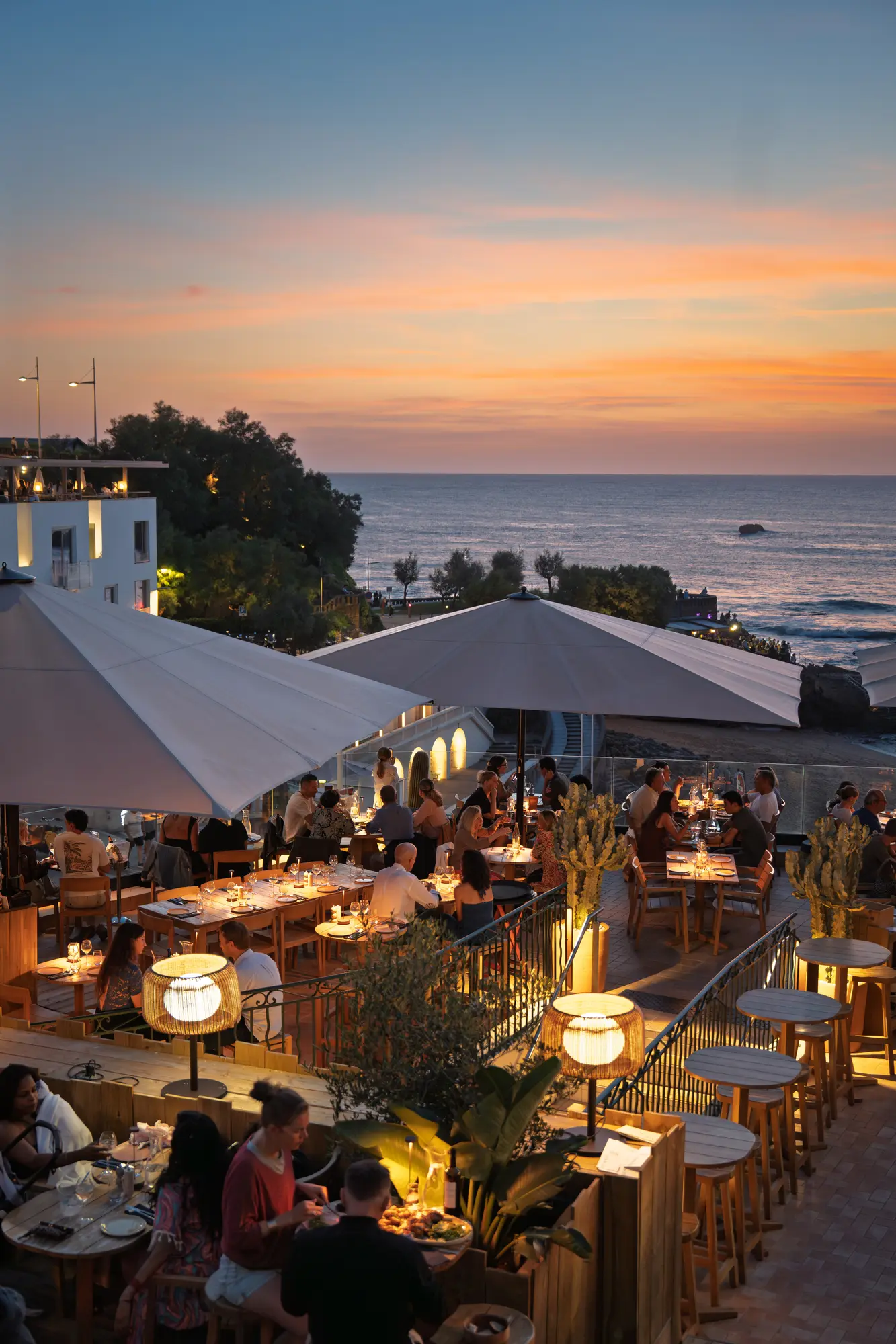 Terrasse de restaurant en soirée au bord de la mer avec des parasols, des tables en bois éclairées par des lampes et des convives dînant avec vue sur un coucher de soleil aux tons orangés et violets.