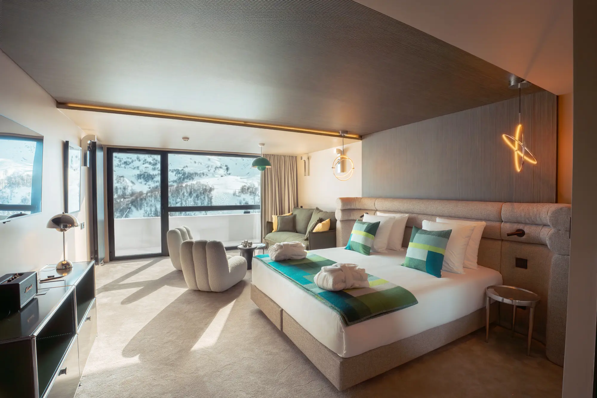 Chambre d'hôtel moderne avec un grand lit double décoré de coussins verts et bleus, deux peignoirs pliés, un fauteuil blanc moelleux, un canapé vert, une table basse, un meuble noir avec une lampe de table, et une grande baie vitrée donnant sur un paysage enneigé de montagnes.