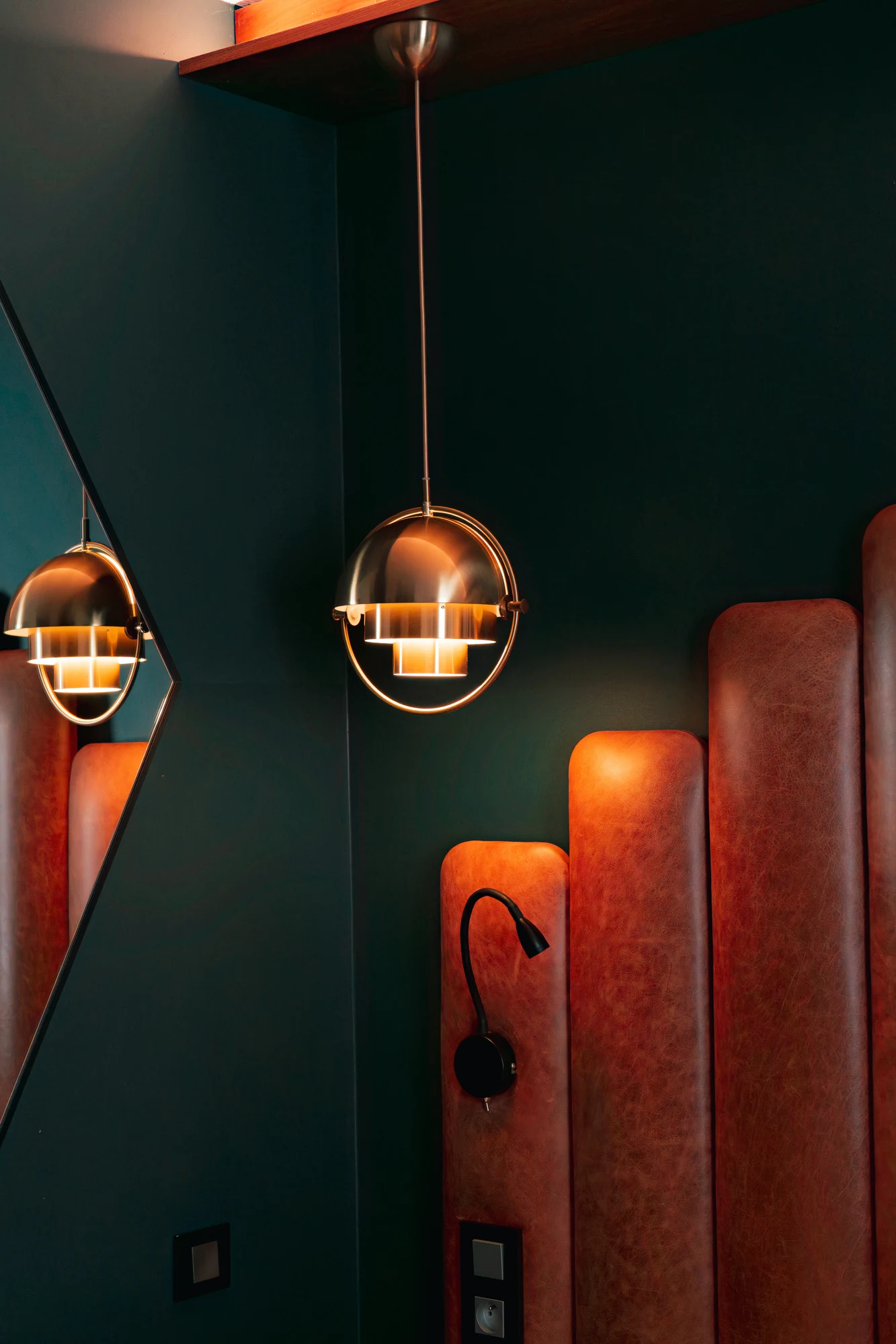 Lampe suspendue métallique dorée avec lumière chaude, reflets dans un miroir à côté d'une tête de lit rembourrée en cuir marron avec liseuse noire montée au mur.