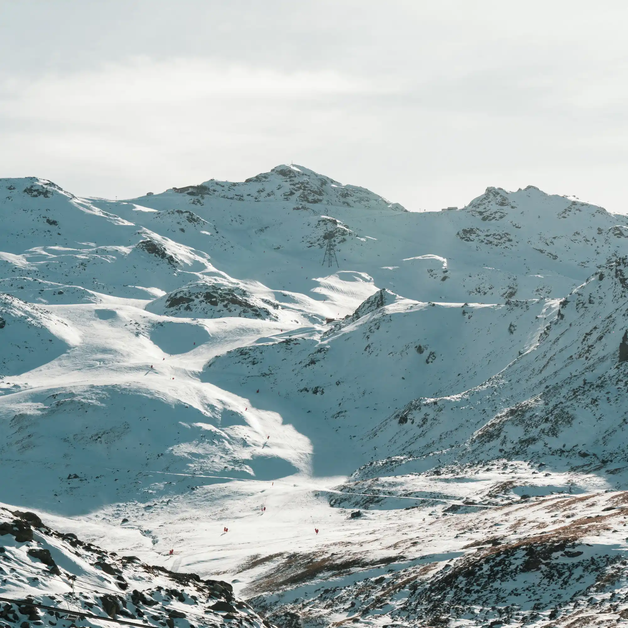 Montagnes enneigées avec des pistes de ski balisées par des drapeaux rouges sous un ciel nuageux clair.