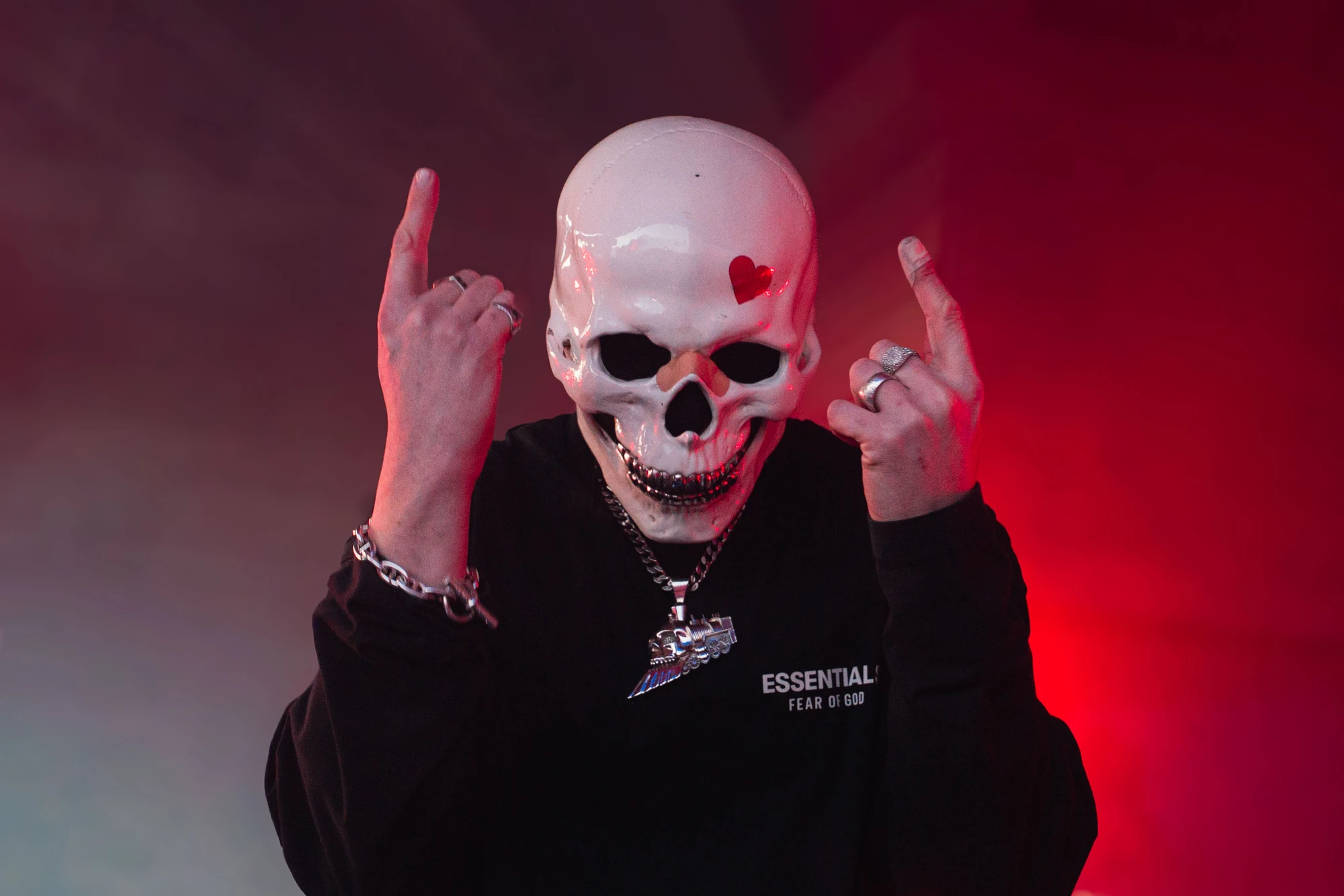 Personne portant un masque de crâne blanc avec un petit cœur rouge sur le front, habillée en noir avec un sweat-shirt portant l'inscription ESSENTIAL FEAR OF GOD, faisant un geste des deux mains avec les doigts levés dans un éclairage rouge et violet.