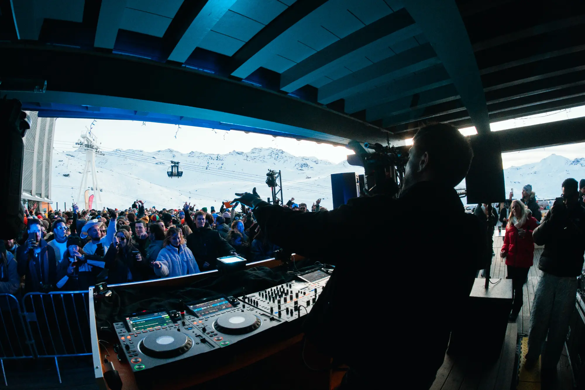 Un DJ joue devant une foule nombreuse sous un abri, avec des montagnes enneigées et une télécabine visibles à l'arrière-plan.
