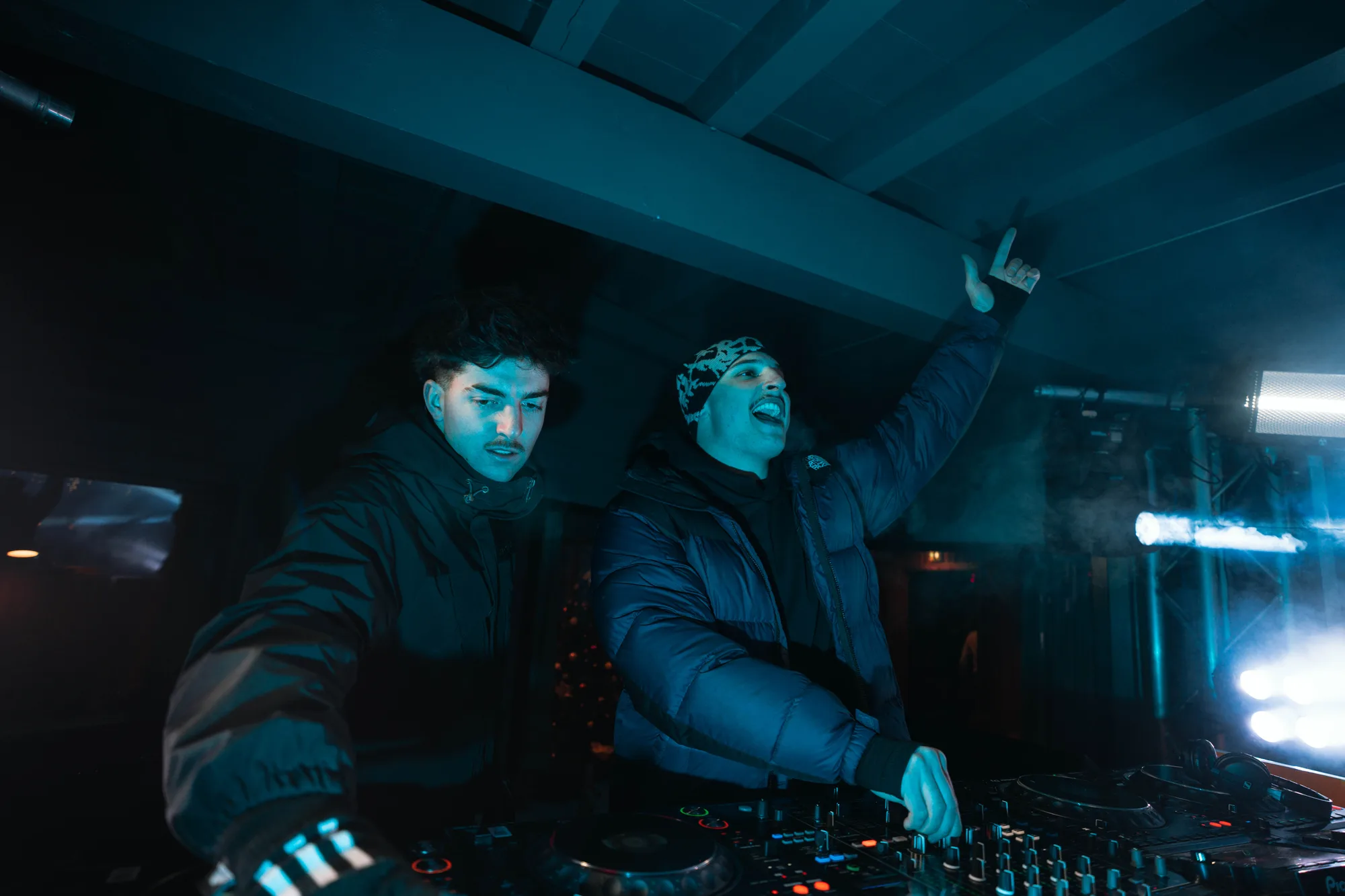 Deux DJs habillés de vestes chaudes sont en train de mixer sur une console dans un environnement sombre avec un éclairage bleu, l'un d'eux levant le bras et souriant.