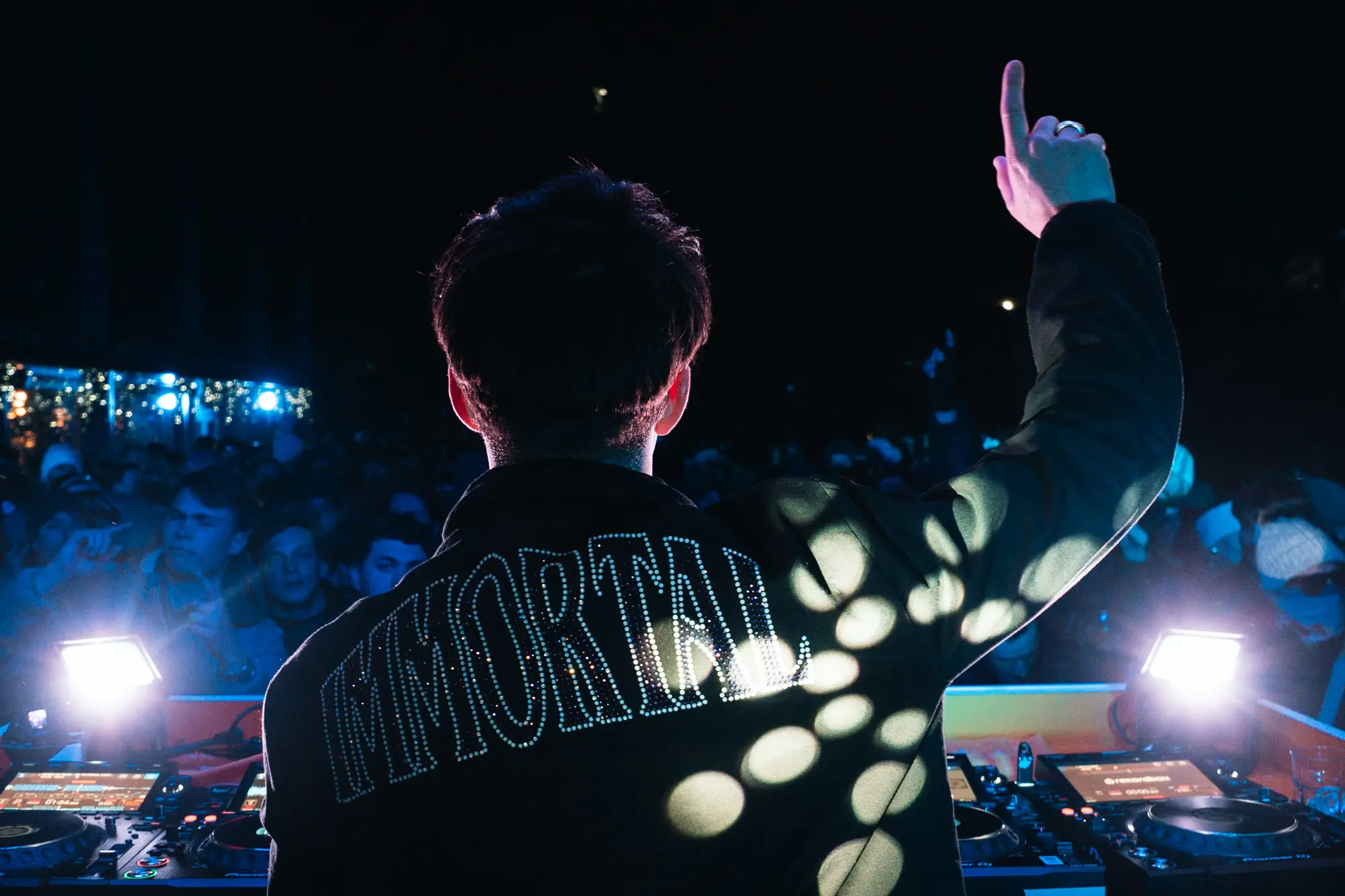 Un DJ vu de dos, levant un doigt vers le ciel dans une boîte de nuit animée, portant une veste noire avec le mot "IMMORTAL" en lettres claires, illuminé par des lumières et des motifs de spots.