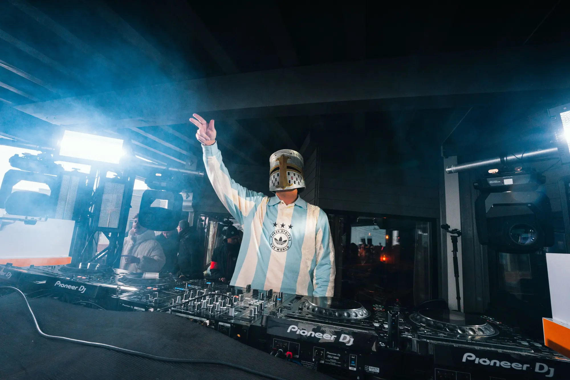 Un DJ portant un casque de chevalier médiéval anime une soirée dans un club sombre avec des équipements Pioneer DJ et des lumières vives et de la fumée en arrière-plan.