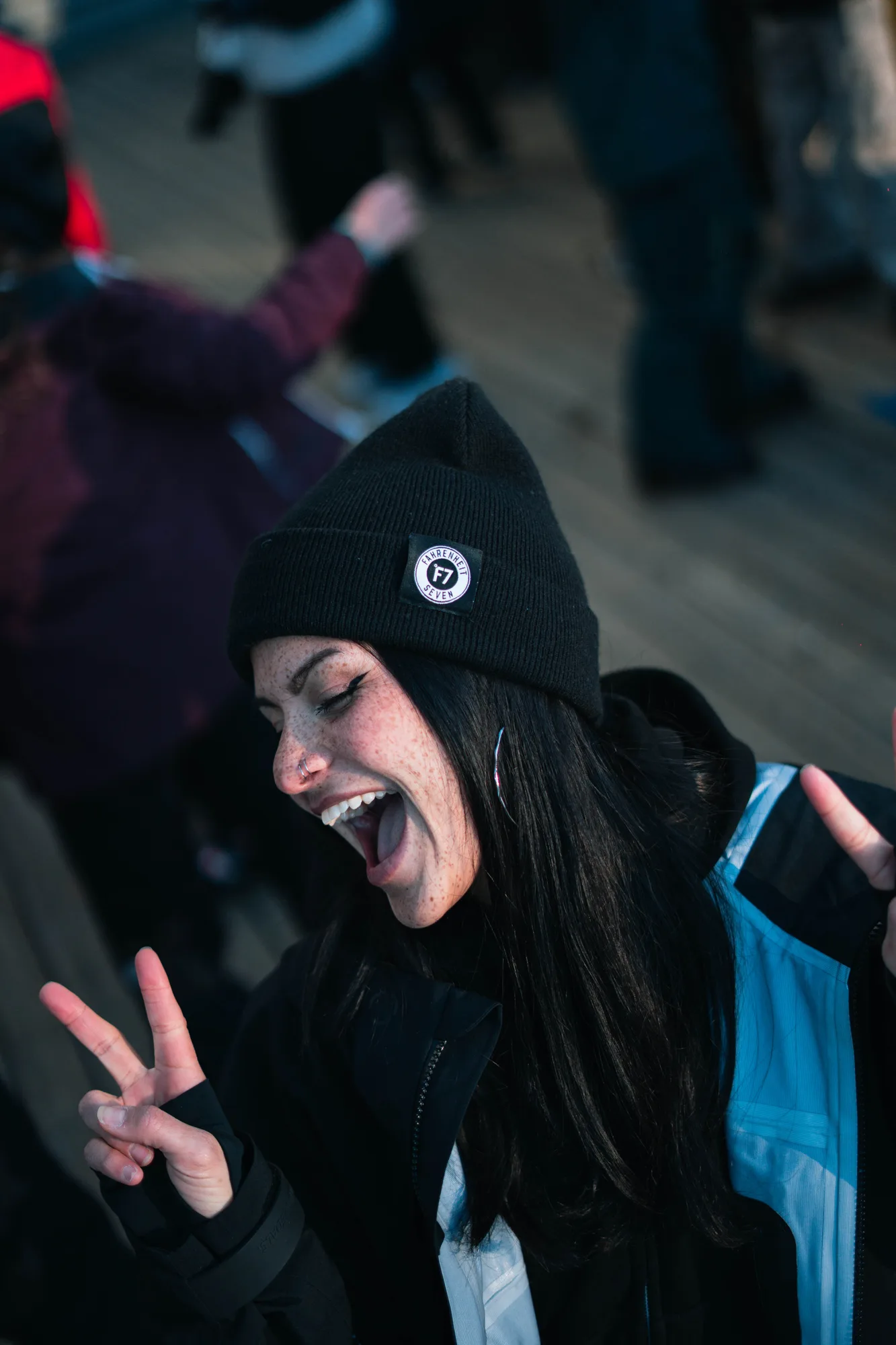 Jeune femme souriante portant un bonnet noir et une veste d'hiver bleu et noire, faisant un signe de paix avec les deux mains et semblant joyeuse dans un environnement extérieur avec d'autres personnes floues en arrière-plan.
