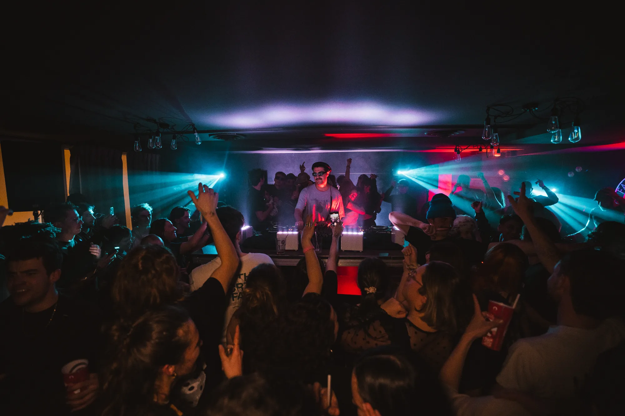 Une foule de personnes dansant et levant les mains lors d'une soirée en intérieur avec un DJ au centre, éclairé par des lumières colorées et des faisceaux lumineux.