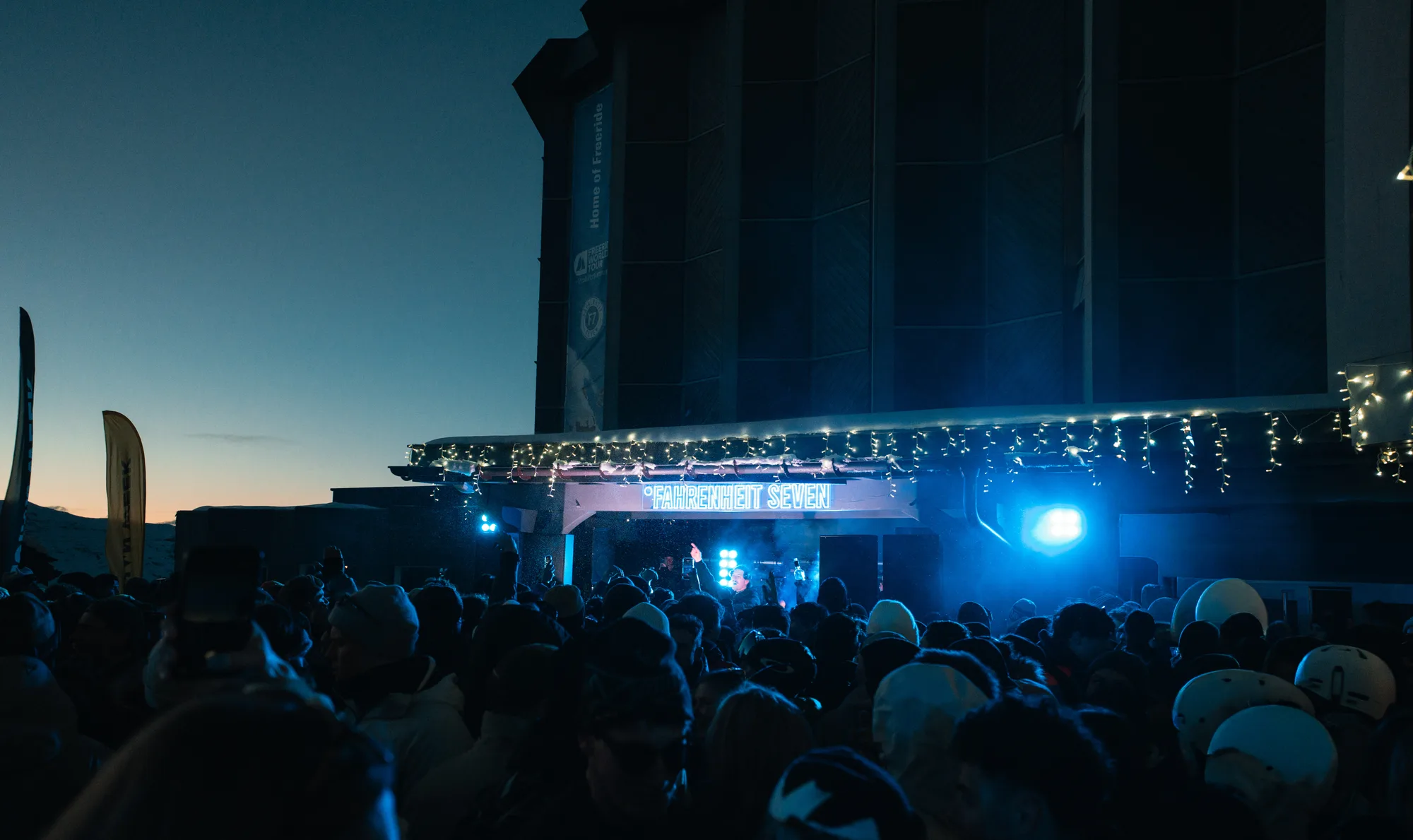 Foule rassemblée de nuit devant une scène illuminée d'un néon affichant "FAHRENHEIT SEVEN", avec des lumières bleues et guirlandes lumineuses décoratives.