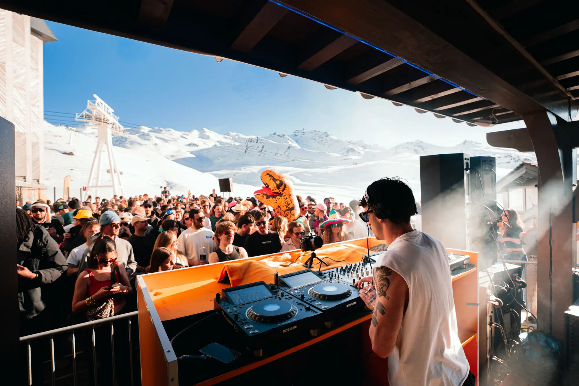 DJ portant un haut sans manche et un casque joue de la musique sur une table de mixage lors d'un concert en plein air en montagne, devant une foule nombreuse sous un ciel bleu.