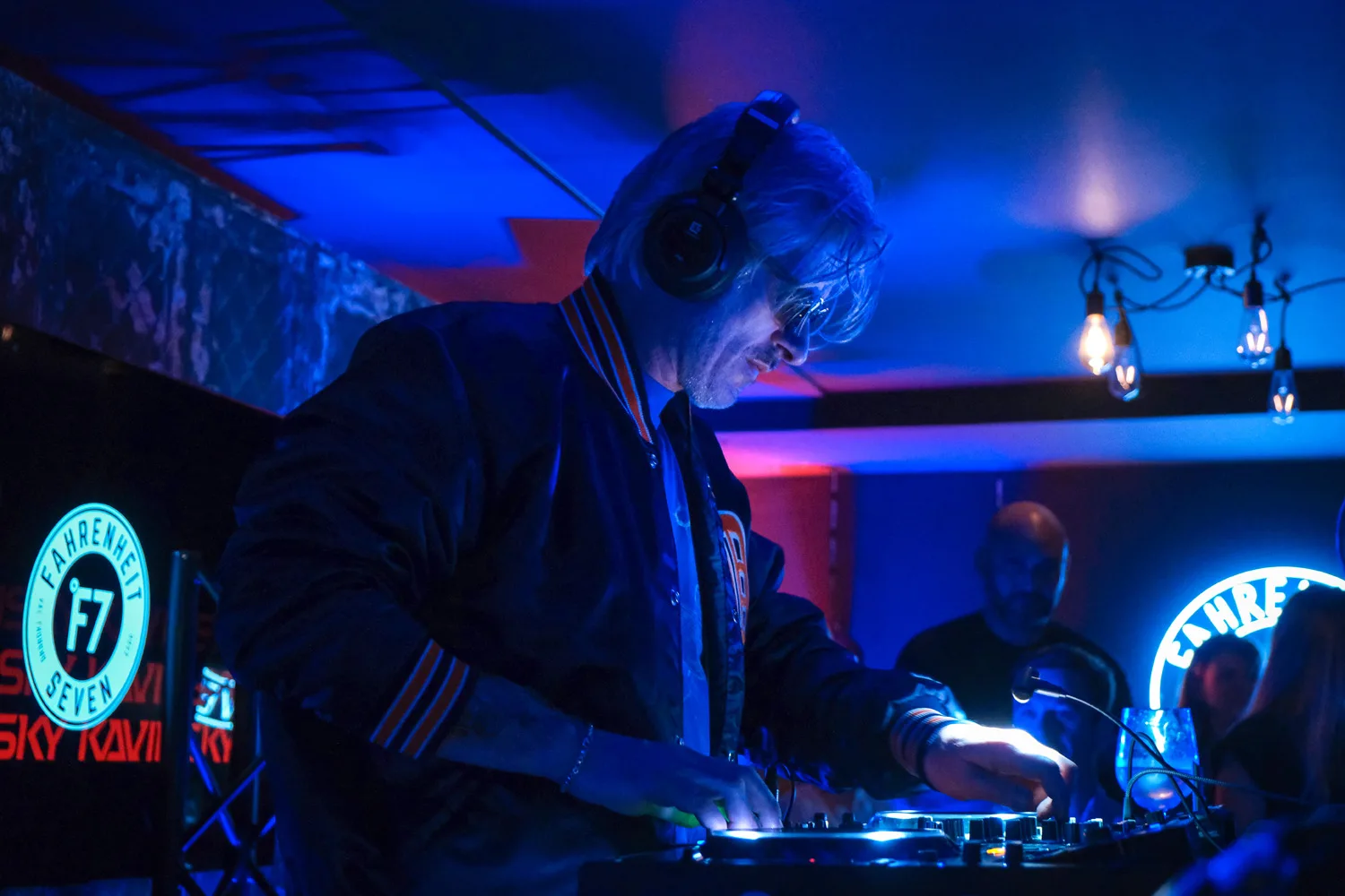 DJ portant un casque et une veste noire aux bandes rouges, mixant de la musique dans un club éclairé en bleu avec un logo lumineux Fahrenheit 7 en arrière-plan.