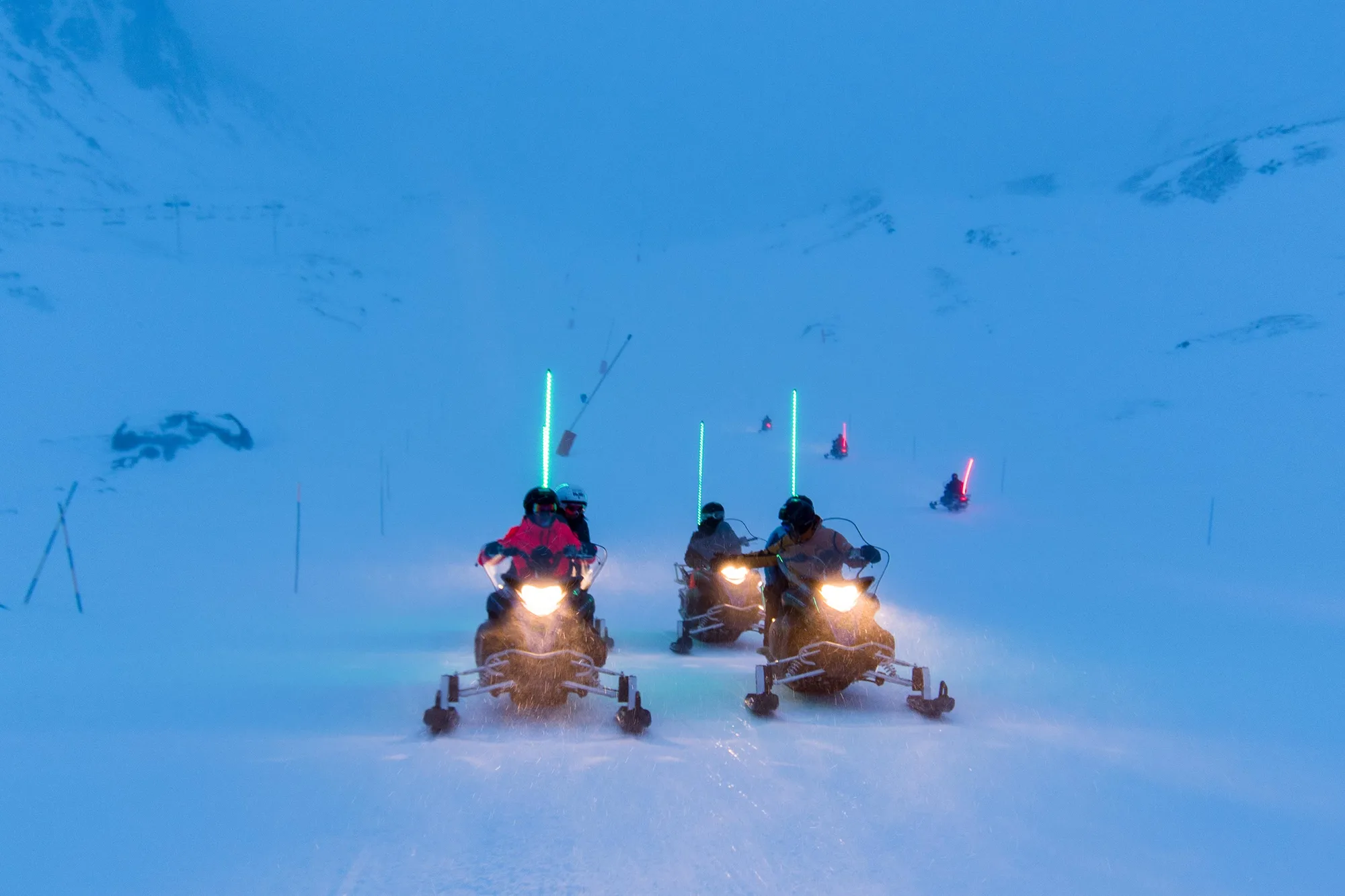 Un groupe de personnes conduisant des motoneiges avec des bâtons lumineux verts et rouges dans un paysage enneigé au crépuscule ou à la tombée de la nuit.