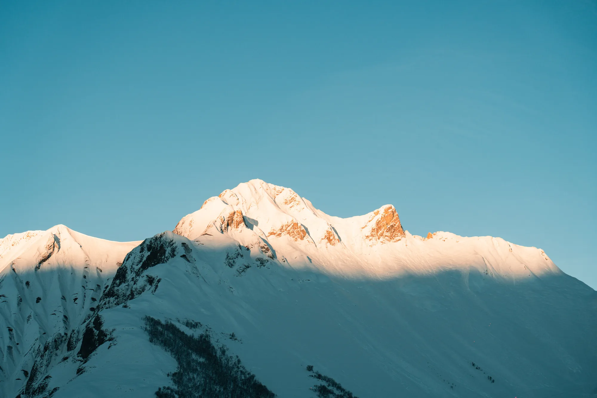 Une montagne enneigée baignée par la lumière du soleil au lever ou coucher du soleil, sous un ciel bleu clair.